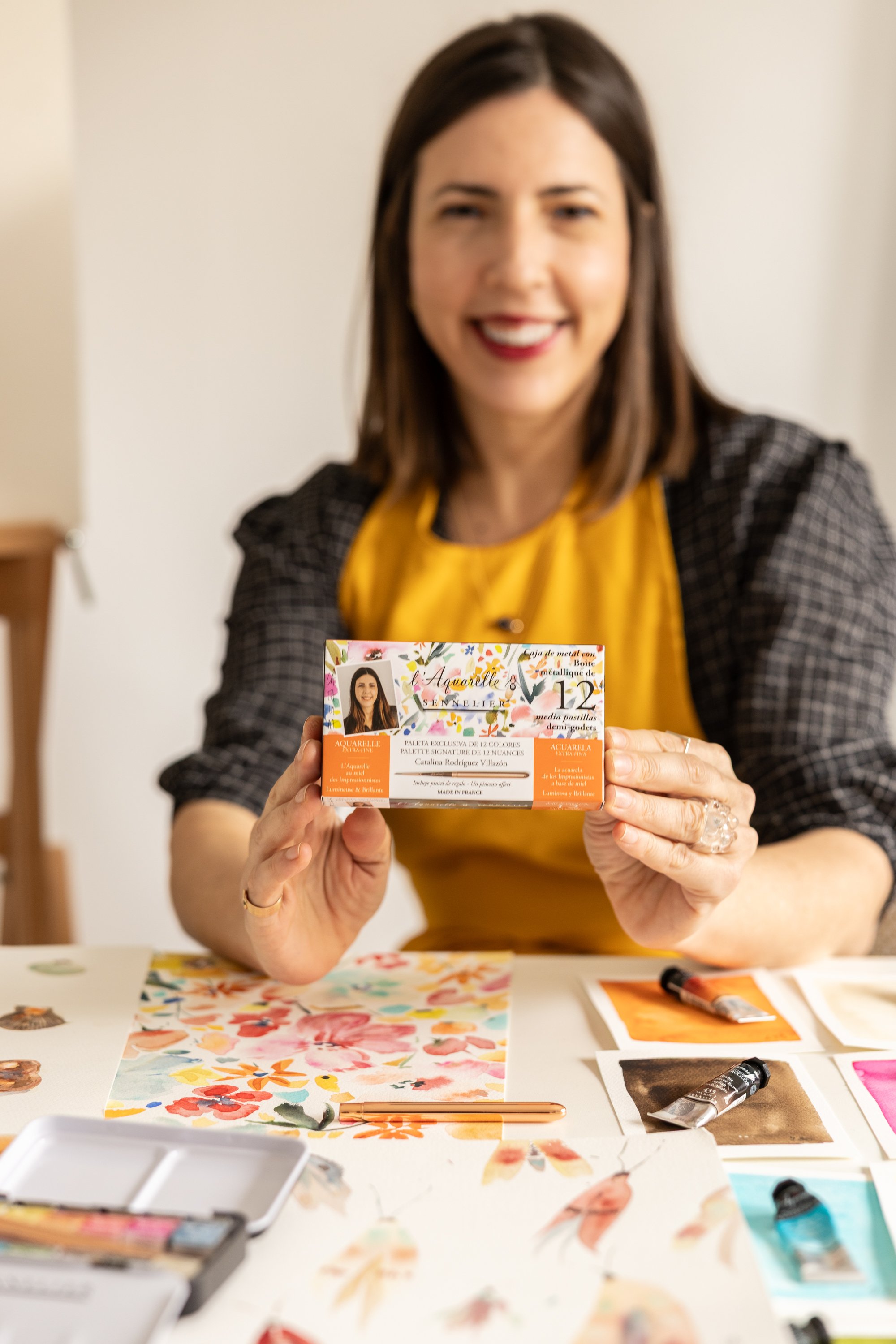 Mujer sonriendo sostiene una caja de acuarelas con flores y colores, en un entorno de trabajo con materiales de arte y papel pintado con acuarela, en un ambiente luminoso.