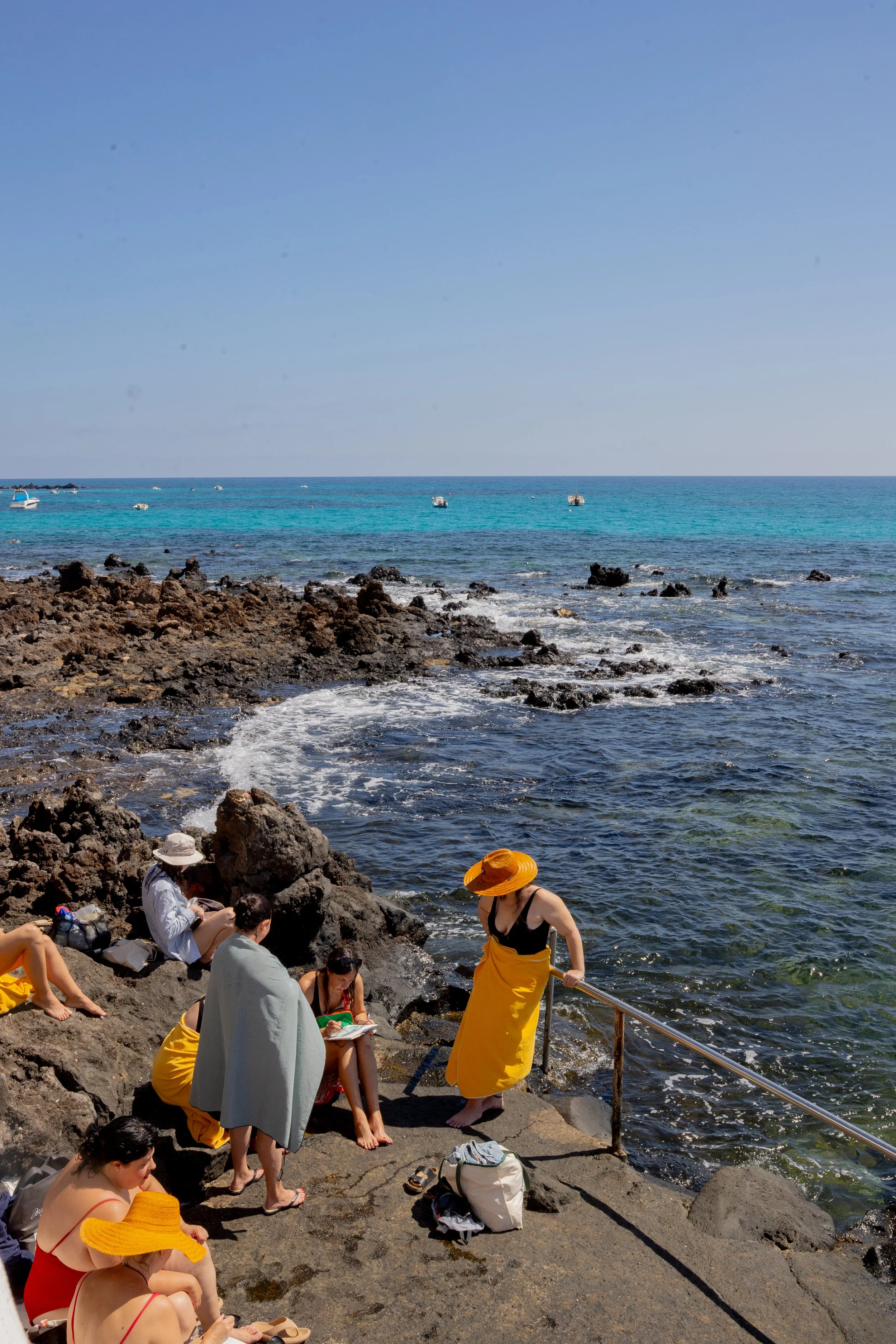 Personas en la playa sentadas en rocas y usando sombreros de ala ancha, con agua y barcos en el mar al fondo.