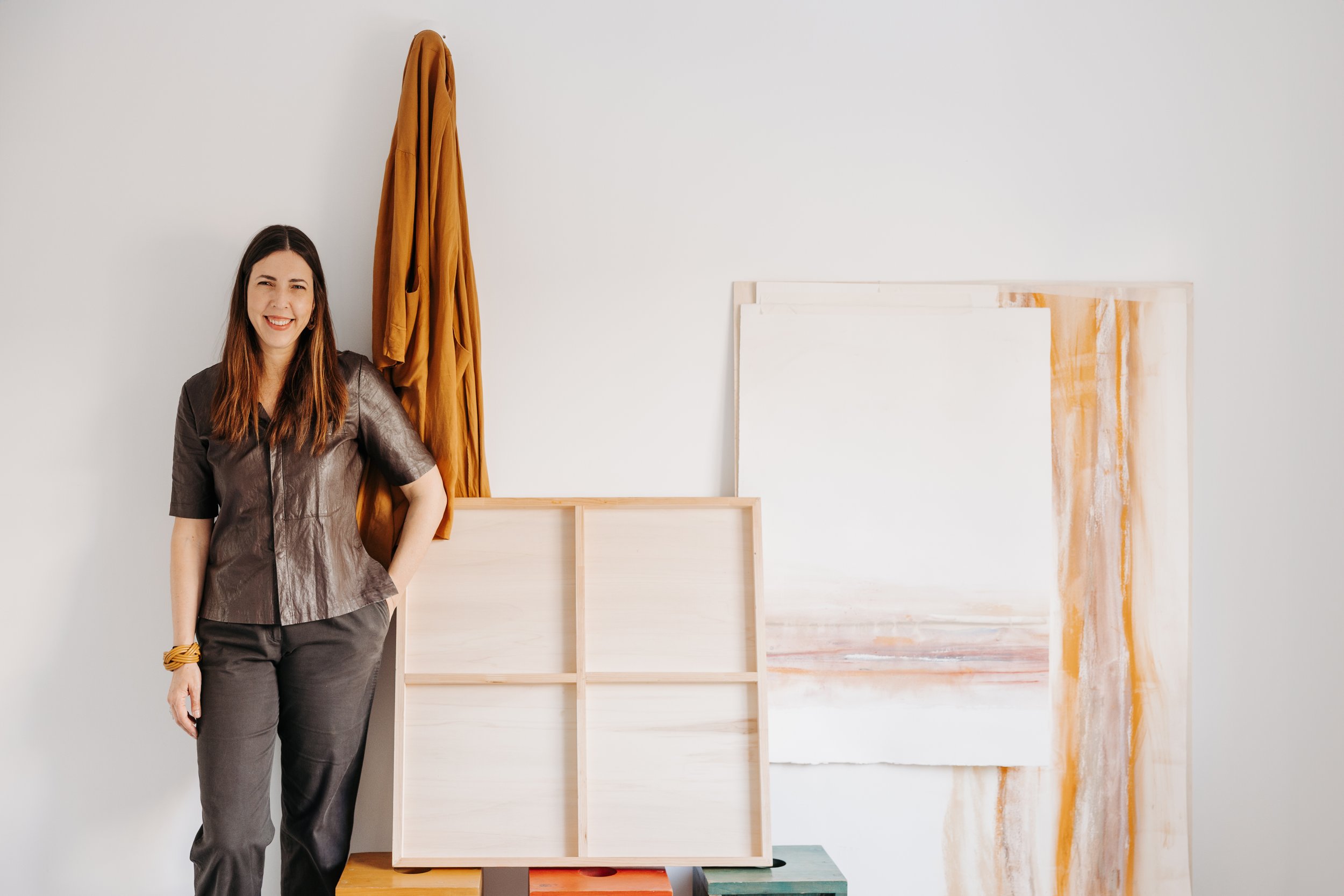 Mujer con cabello largo, de camiseta gris y pantalones oscuros, sonriendo, frente a una pared blanca con varios lienzos y un lienzo grande de pintura abstracta en tonos blancos, naranjas y dorados.