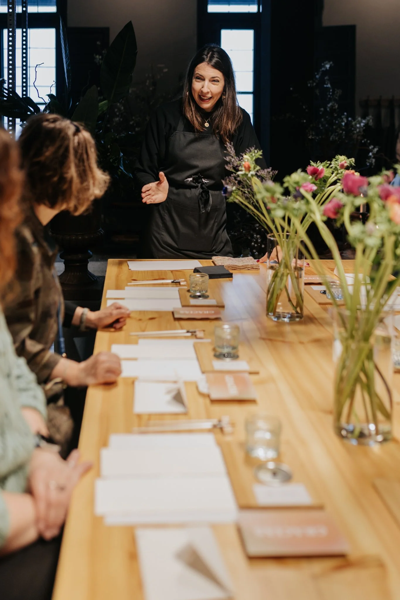 Mujer de cabello castaño y ropa negra, sonriendo y explicando algo a un grupo sentado en una mesa de madera rodeada de flores y documentos.