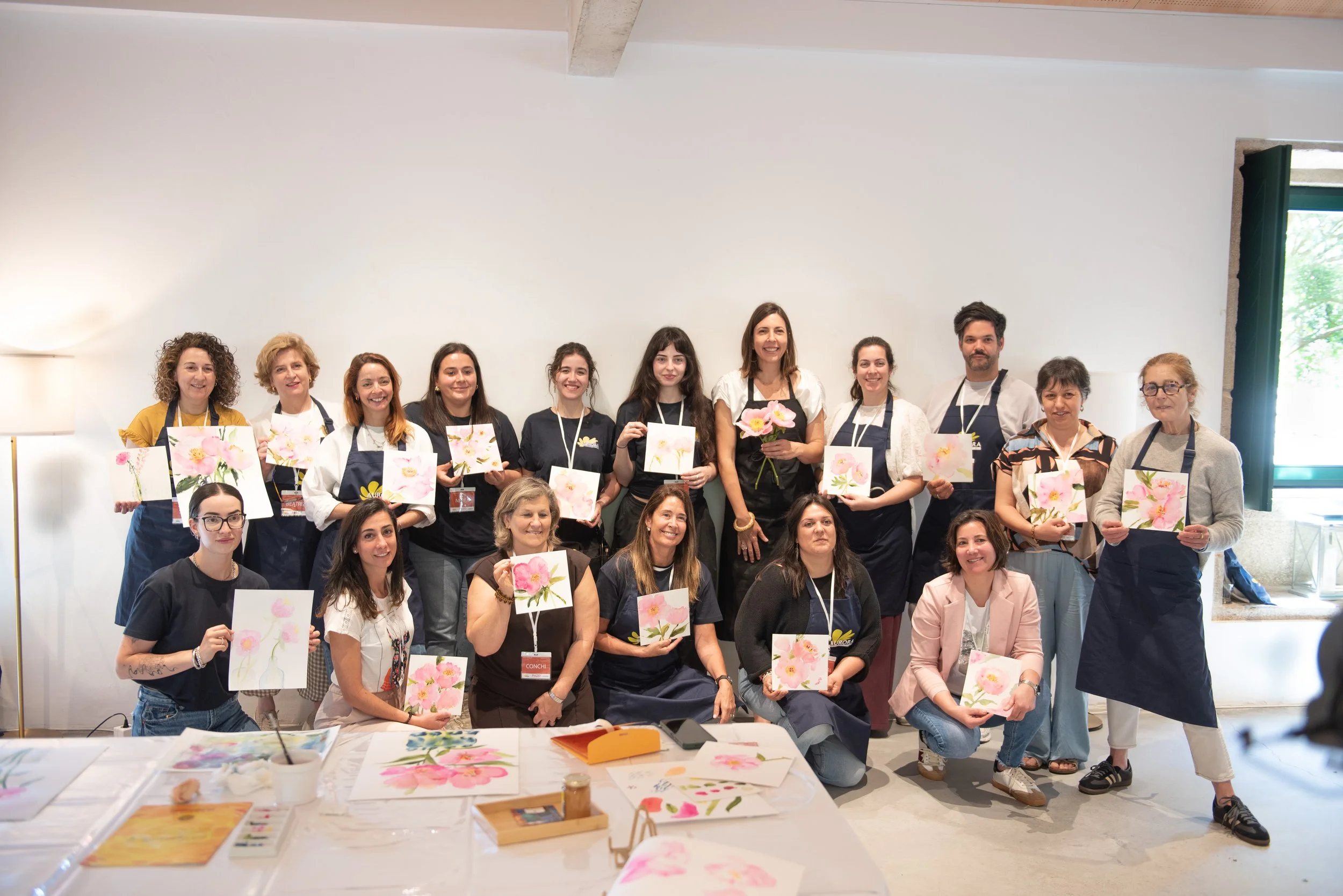 Grupo de personas en un taller de pintura, sosteniendo obras con flores en acuarela y un florero con flores reales, en un espacio interior con luz natural.