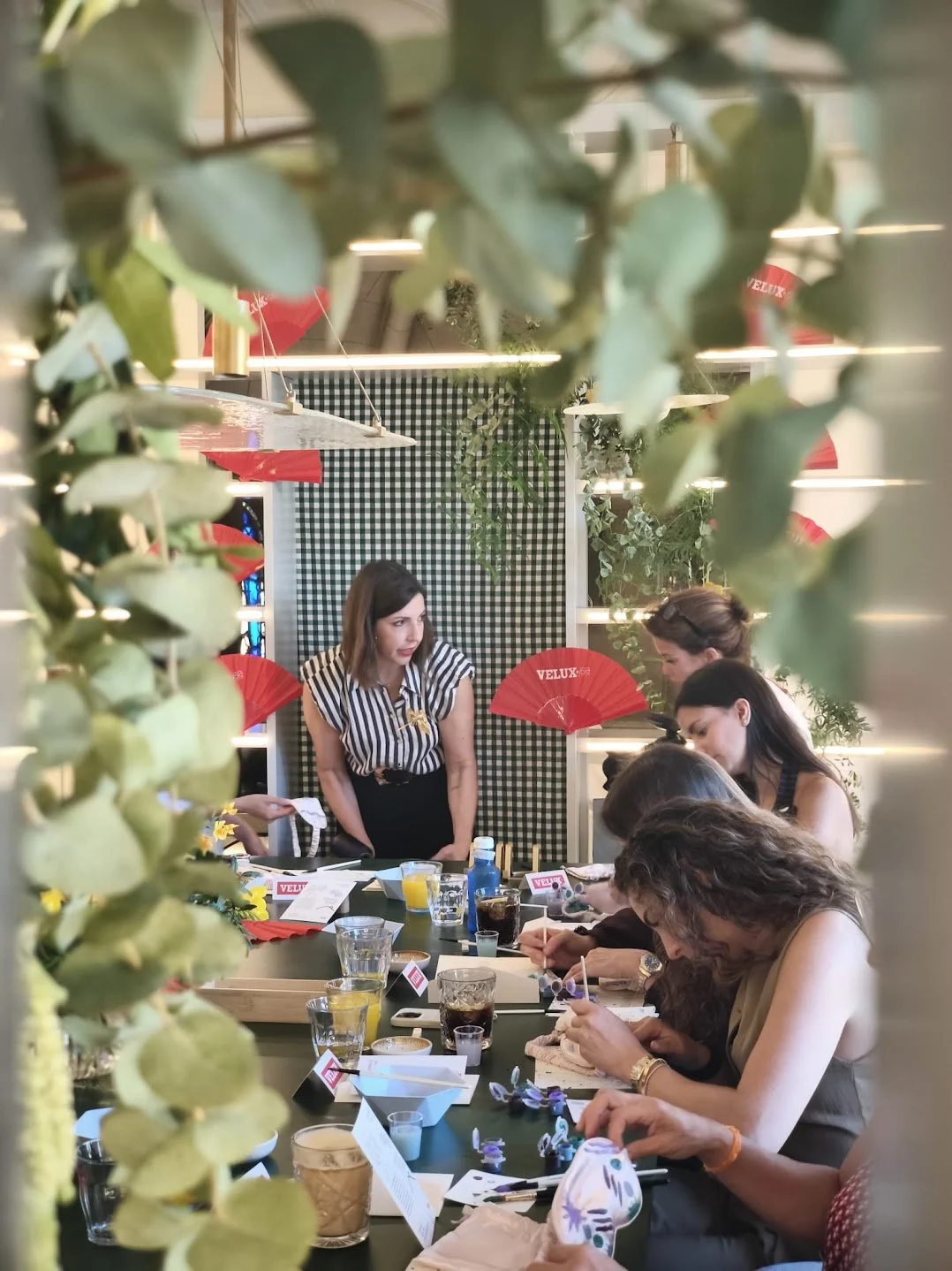 Grupo de personas en una reunión o taller en un espacio decorado con fanales rojos y plantas verdes, vista a través de hojas en primer plano.