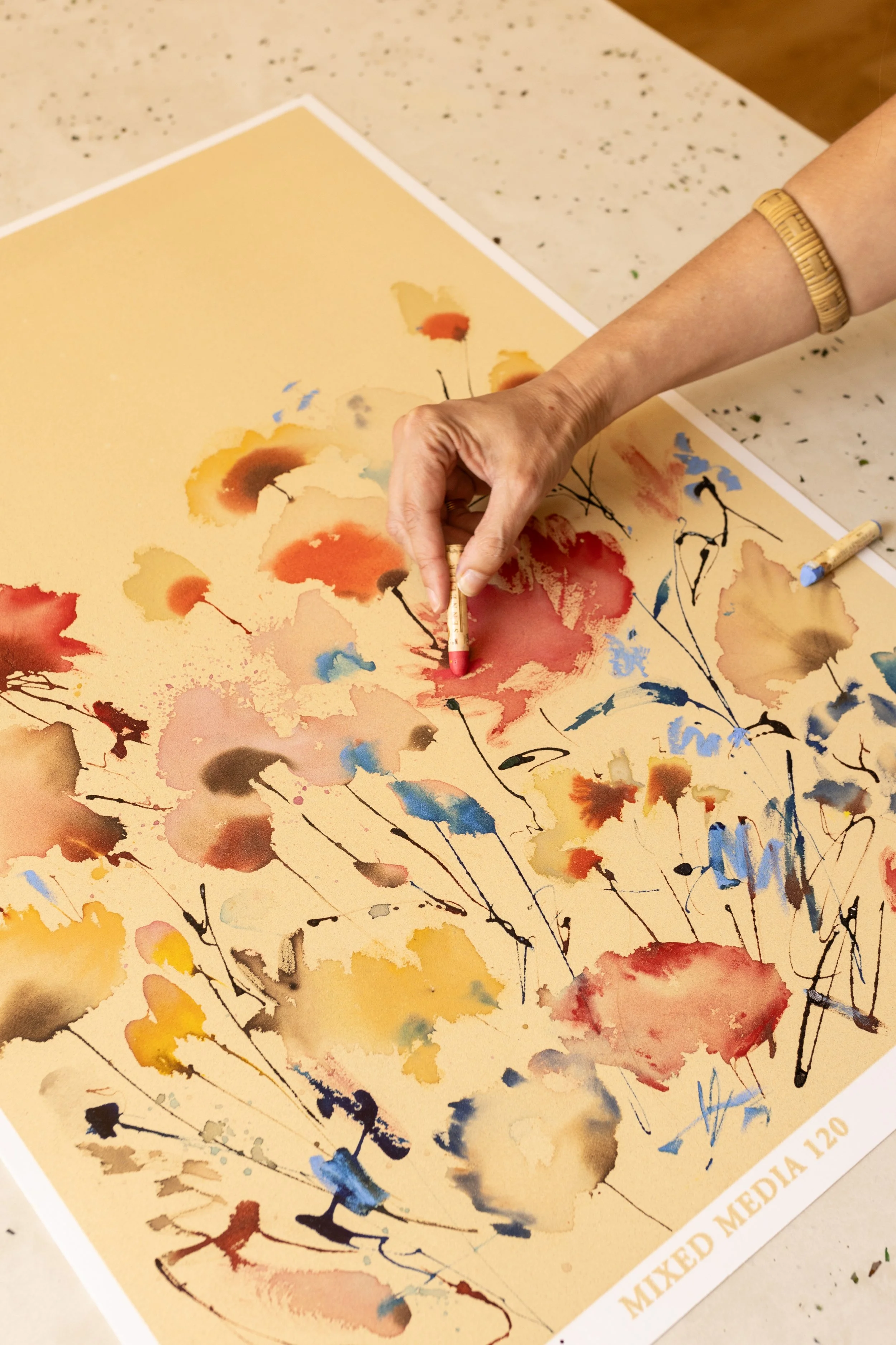 Mano con crayón pintando un dibujo acuarelado de flores en un papel color beige en una mesa.