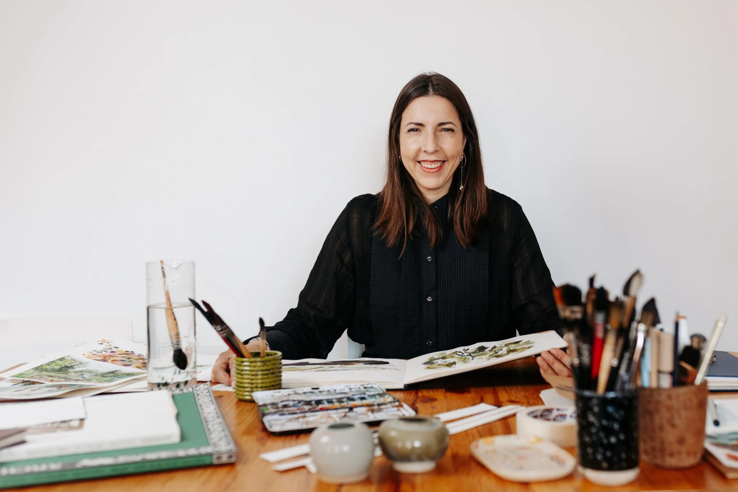 Mujer sonriente en una mesa de arte, rodeada de libros, cerámicas y materiales de pintura.