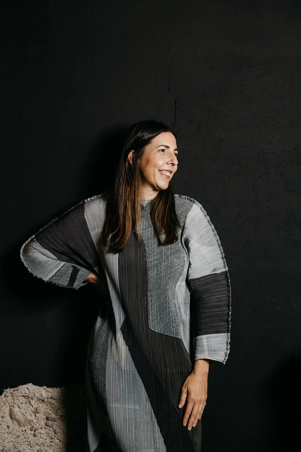 Mujer sonriendo, de cabello largo y oscuro, usando un vestido de patrones en tonos gris, negro y blanco, de fondo negro.