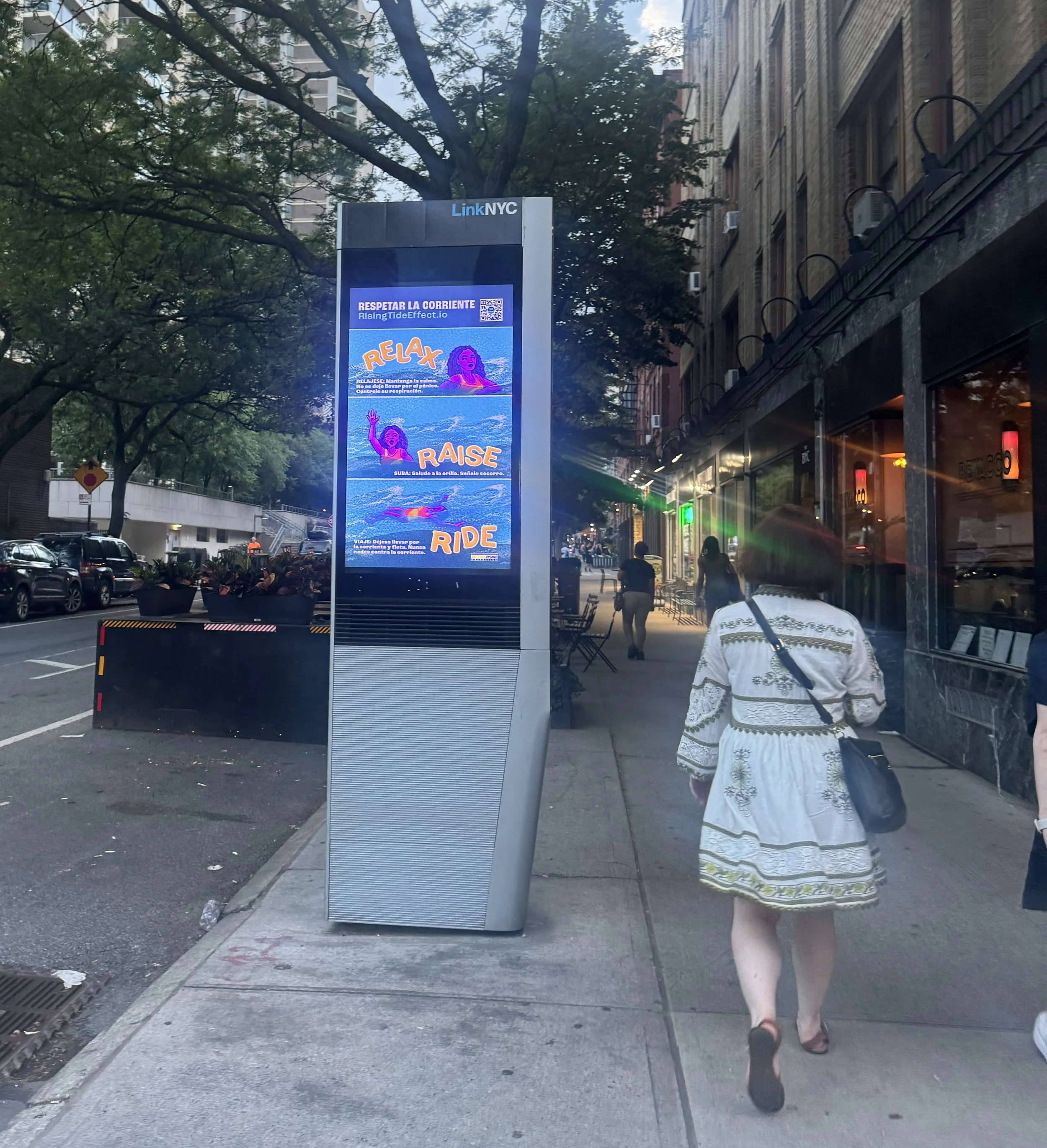 A digital outdoor billboard on a city sidewalk displaying a message about water safety with illustrations of a girl swimming and the words "Relax," "Raise," and "Ride."