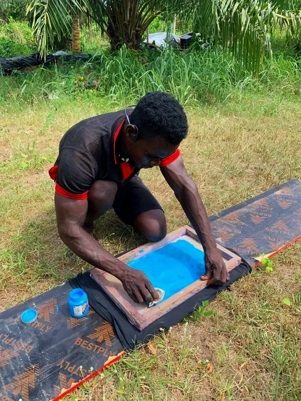 A man kneeling outdoors on grass, using a sponge to create blue ink on a screen printing frame. He is surrounded by trees and greenery, with some black material and equipment in the background.
