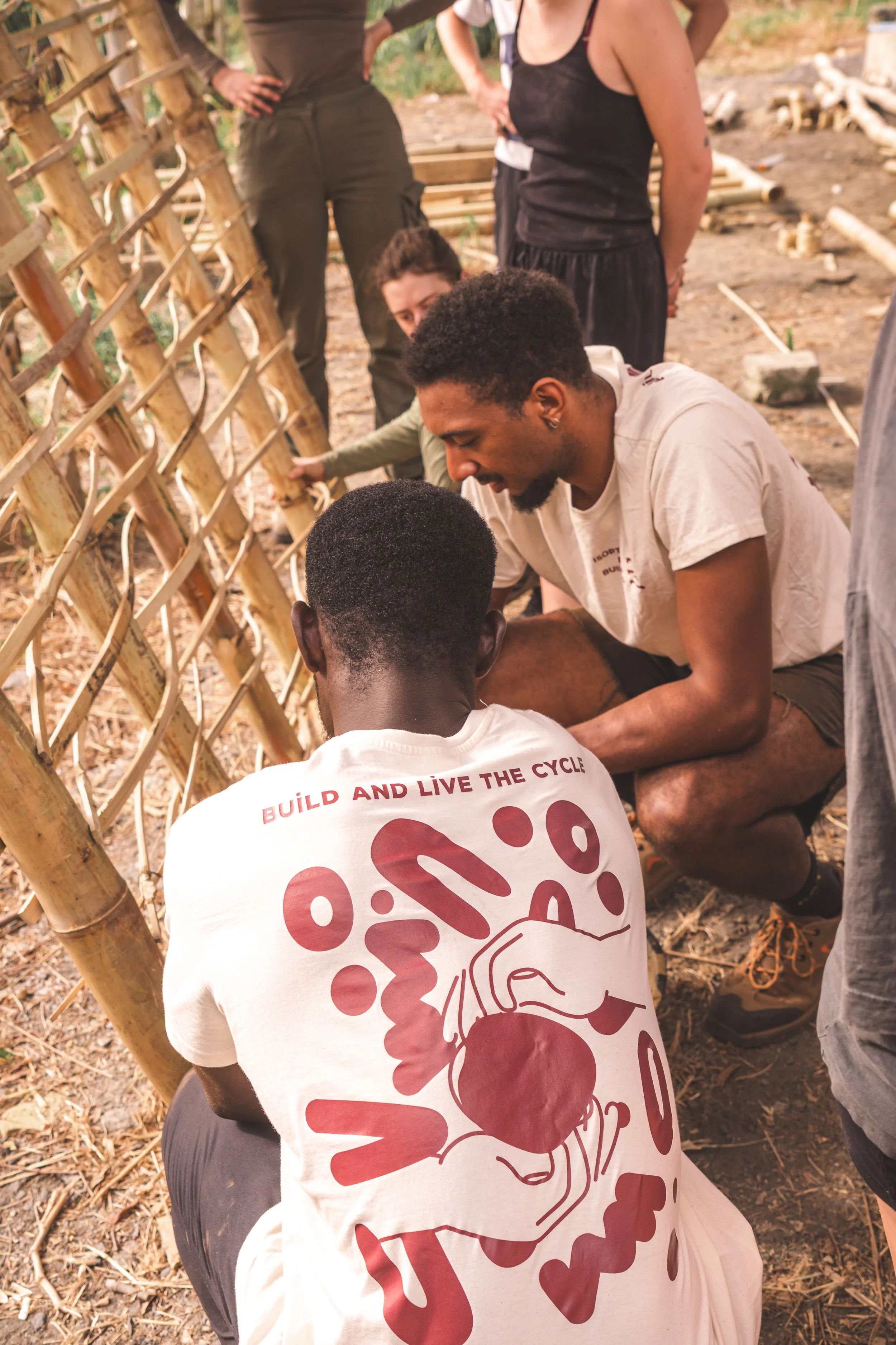 Group of people working together to build a bamboo structure outdoors on a dirt ground, with some logs and construction materials scattered around.