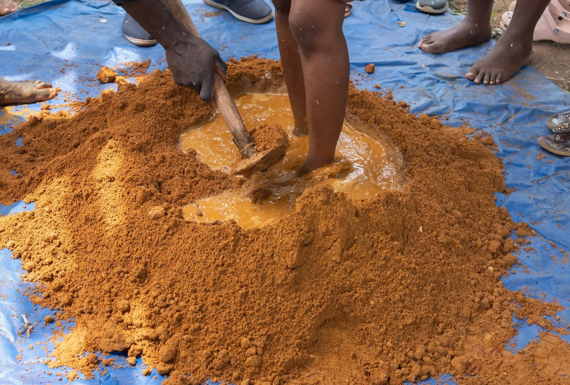 People standing around a blue plastic sheet with a mound of reddish-brown mud or clay in the center. One person is mixing or working with the mud using a wooden stick.