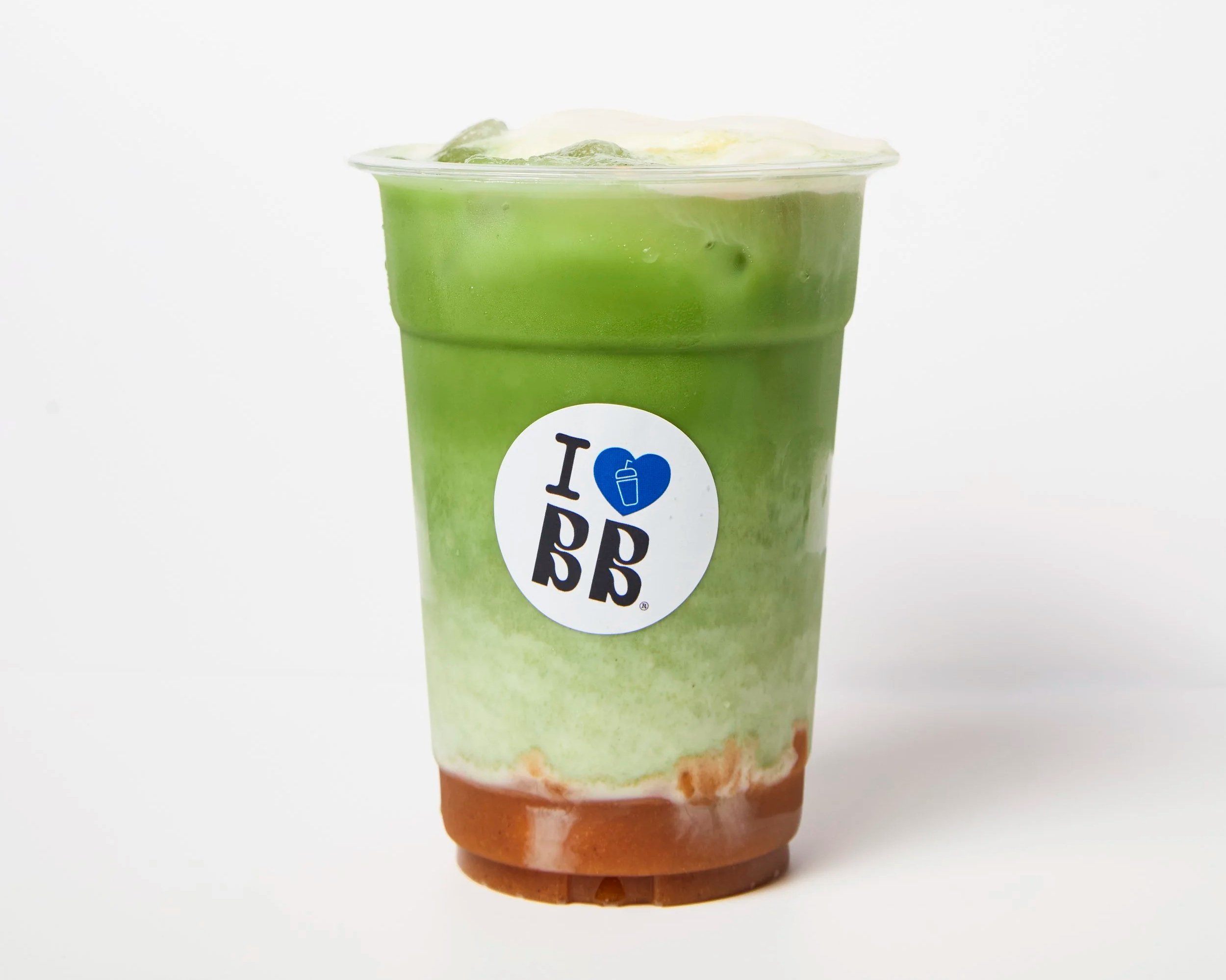 A cup of green layered frozen beverage with colorful swirls, topped with a thin layer of white foam, and a brown base. The cup has a logo with the text "I ♥ BP".