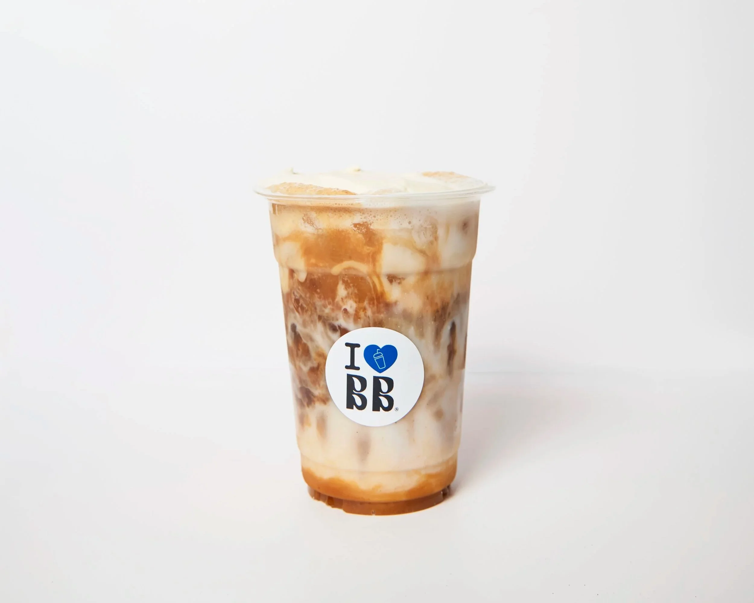 A cold beverage in a clear plastic cup with ice, caramel syrup, and milk, topped with whipped cream, on a white background.