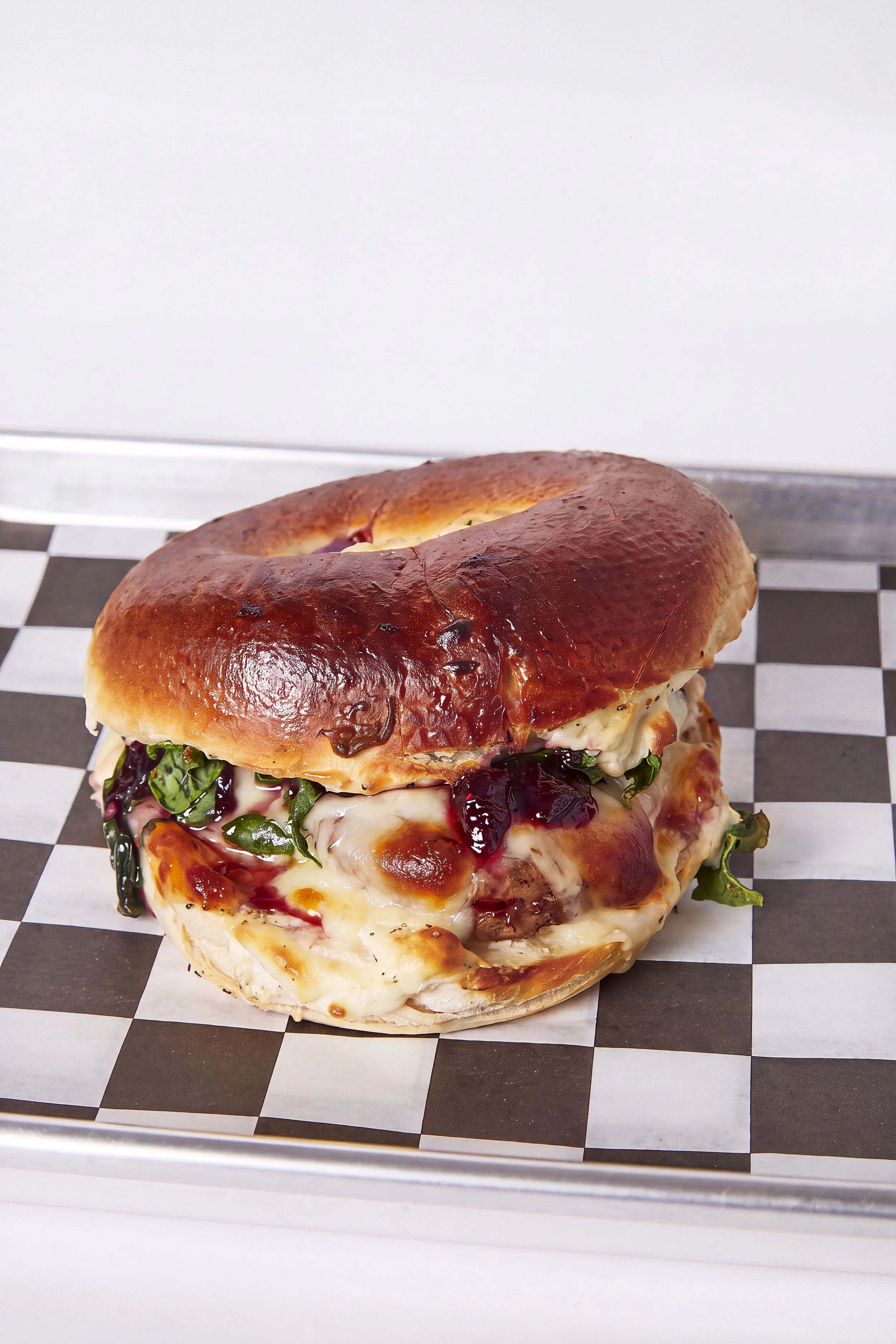 A baked chicken sandwich with cheese, lettuce, tomato, and a dark sauce on a checkered paper surface.