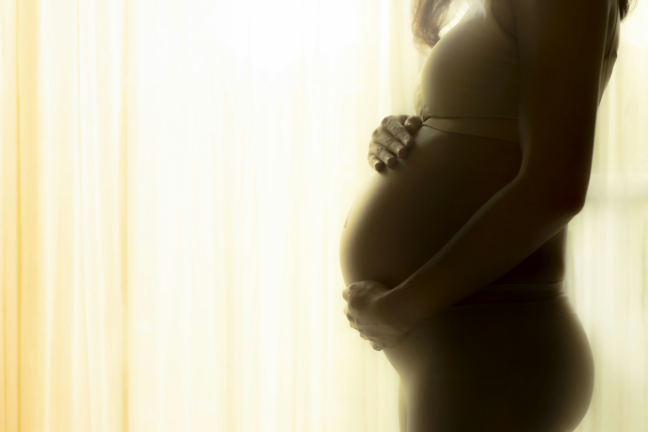 Silhouette of a woman with long hair, standing with one hand on her pregnant belly near a window with yellow curtains.