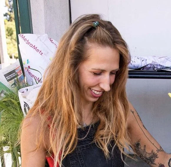 A young woman with long, wavy blonde hair, wearing a black top and denim overalls, smiling and looking down.