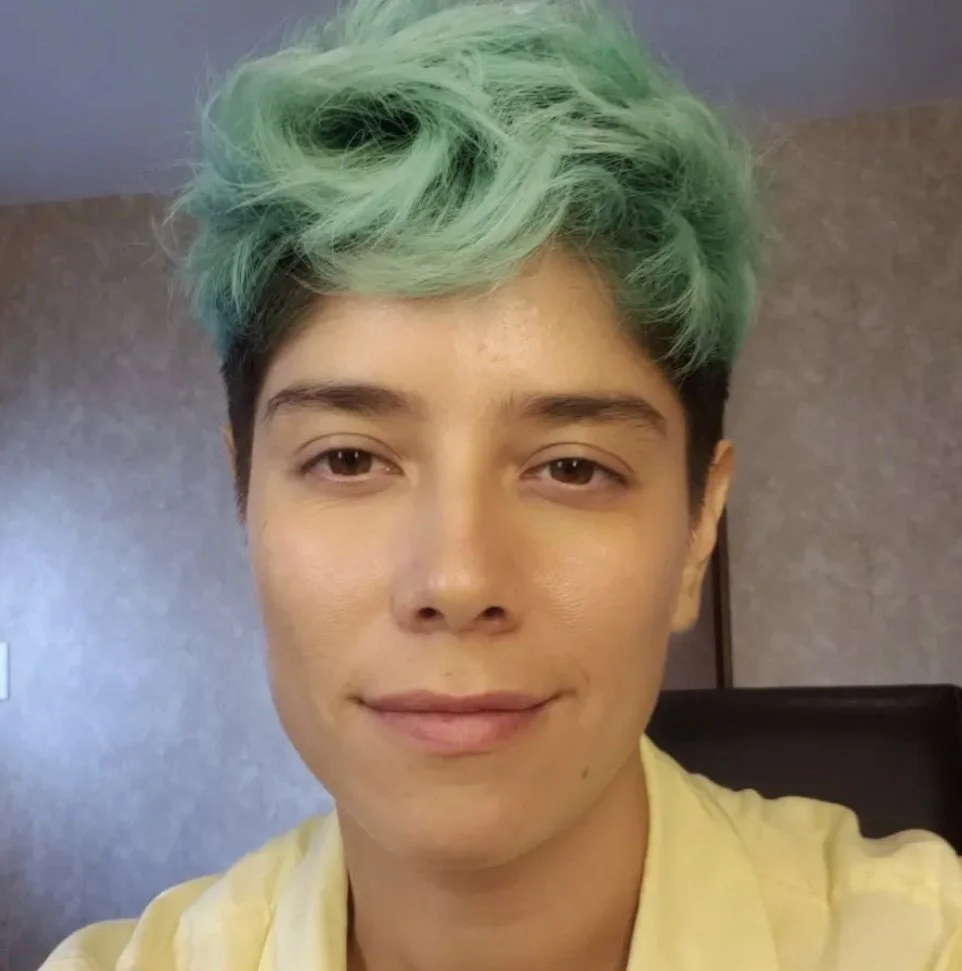 A person with short, black hair and mint green hair on top, smiling and wearing a yellow shirt indoors.