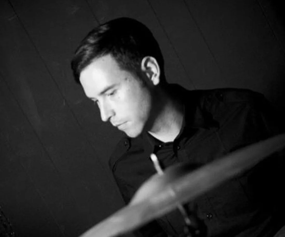 A black and white photo of a young man with slicked-back hair, looking downward and slightly to the side, wearing a dark shirt.