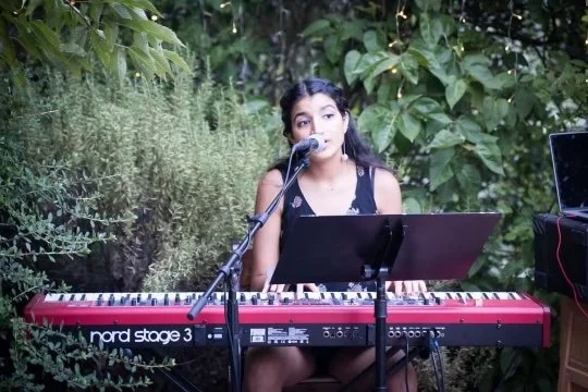 Young woman singing and playing a red Nord Stage 3 keyboard outdoors surrounded by green foliage.