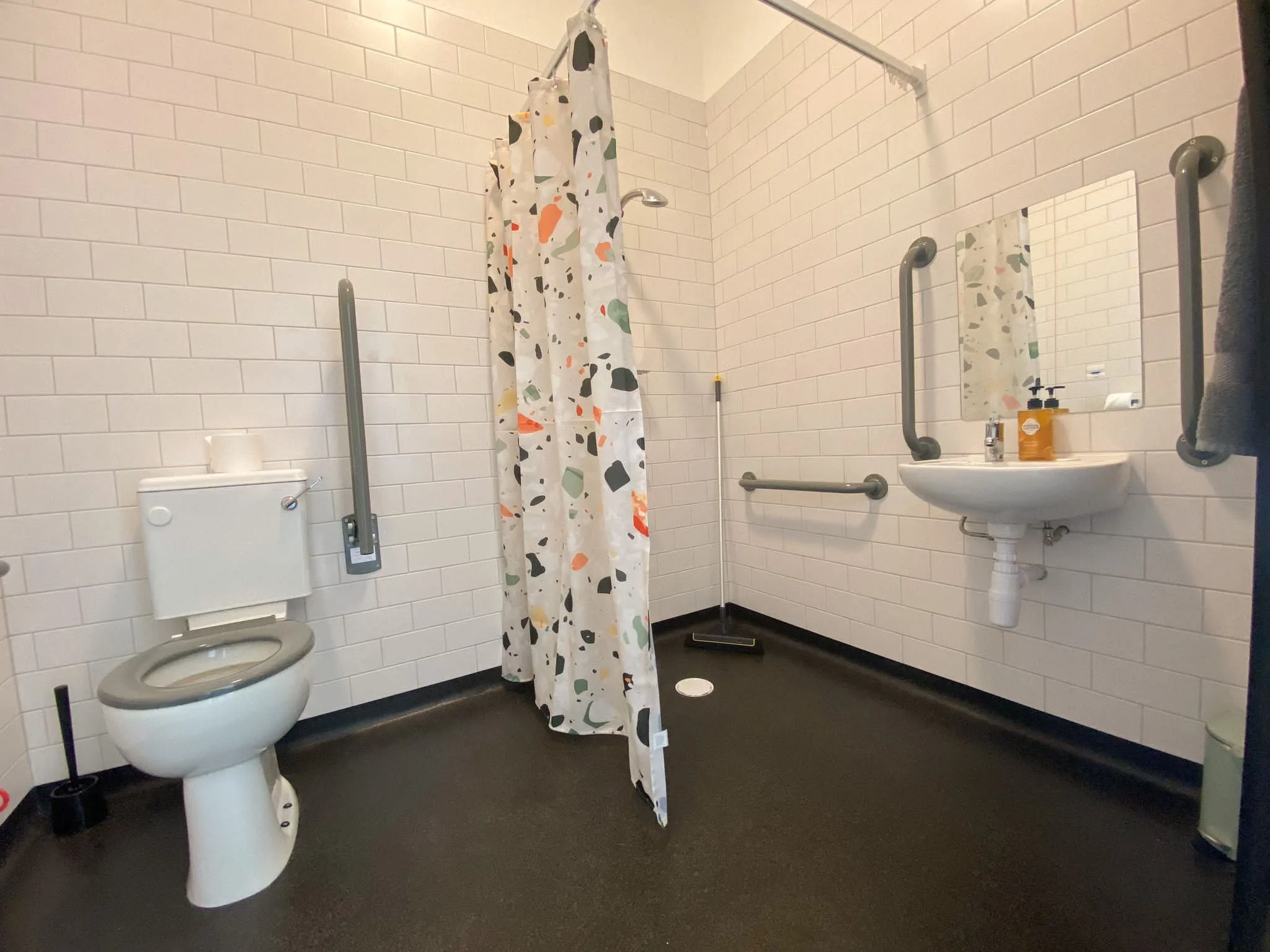 Accessible bathroom with a toilet, a shower area with a colorful shower curtain, grab bars, a small wall-mounted sink, and a mirror.
