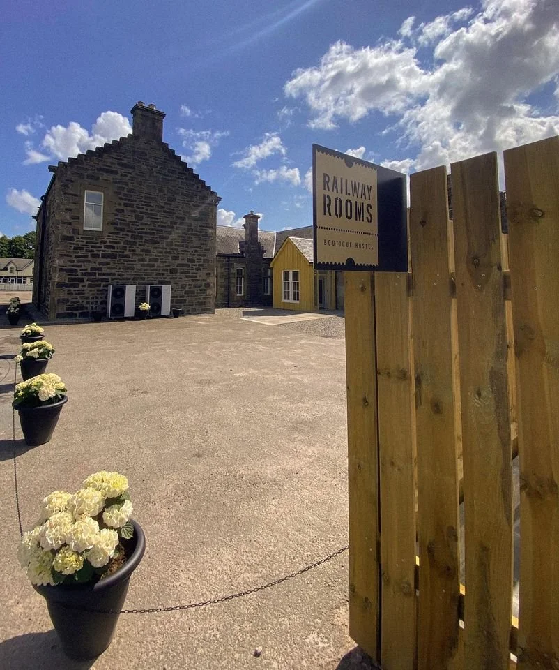 A rustic stone building with a yellow addition in the background, white flowers in black pots along the edge, a sign that reads 'Railway Rooms Boutique Hostel,' and a partly cloudy blue sky.