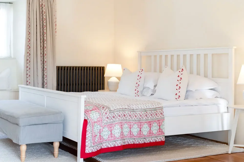 A neatly made bed with white bedding, decorative pillows with floral embroidery, a patterned throw blanket, beside two white nightstands with lamps, in a bright room with curtains and a window.