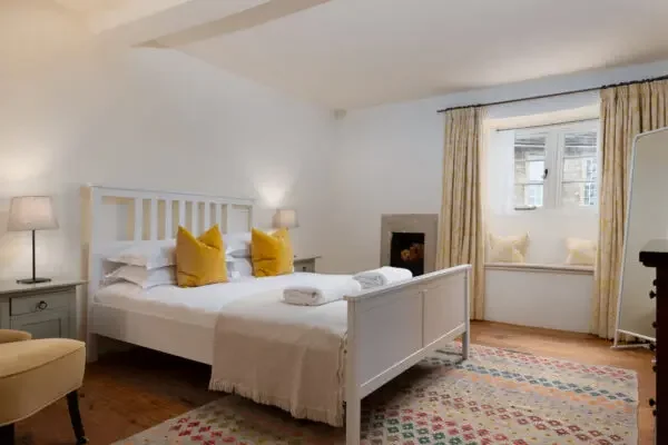 A cozy bedroom with a white bed, yellow pillows, bedside lamps, a window seat with yellow cushions, and a colorful rug.