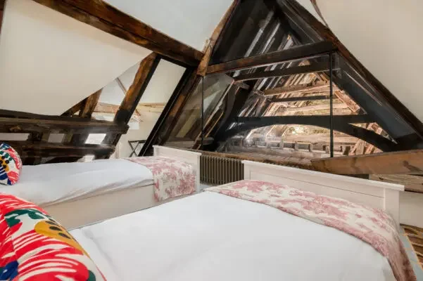 Two beds with white sheets and pink blankets in an attic bedroom with sloped ceiling and exposed wooden beams and windows.