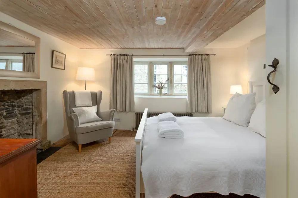 Cozy bedroom with a white bed, rolled towels, wooden ceiling, armchair, small table with lamp, fireplace, and window with curtains.