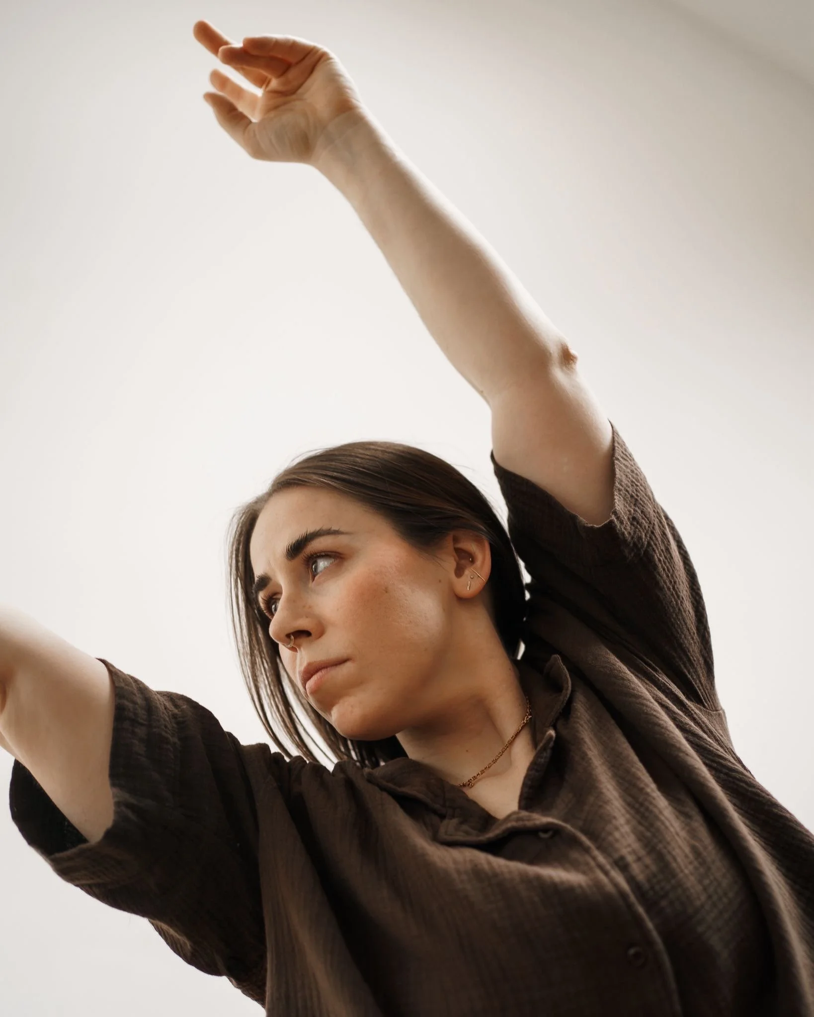 A young woman with brown hair and earrings is raising her arm, with her hand close to the camera, against a plain light background.