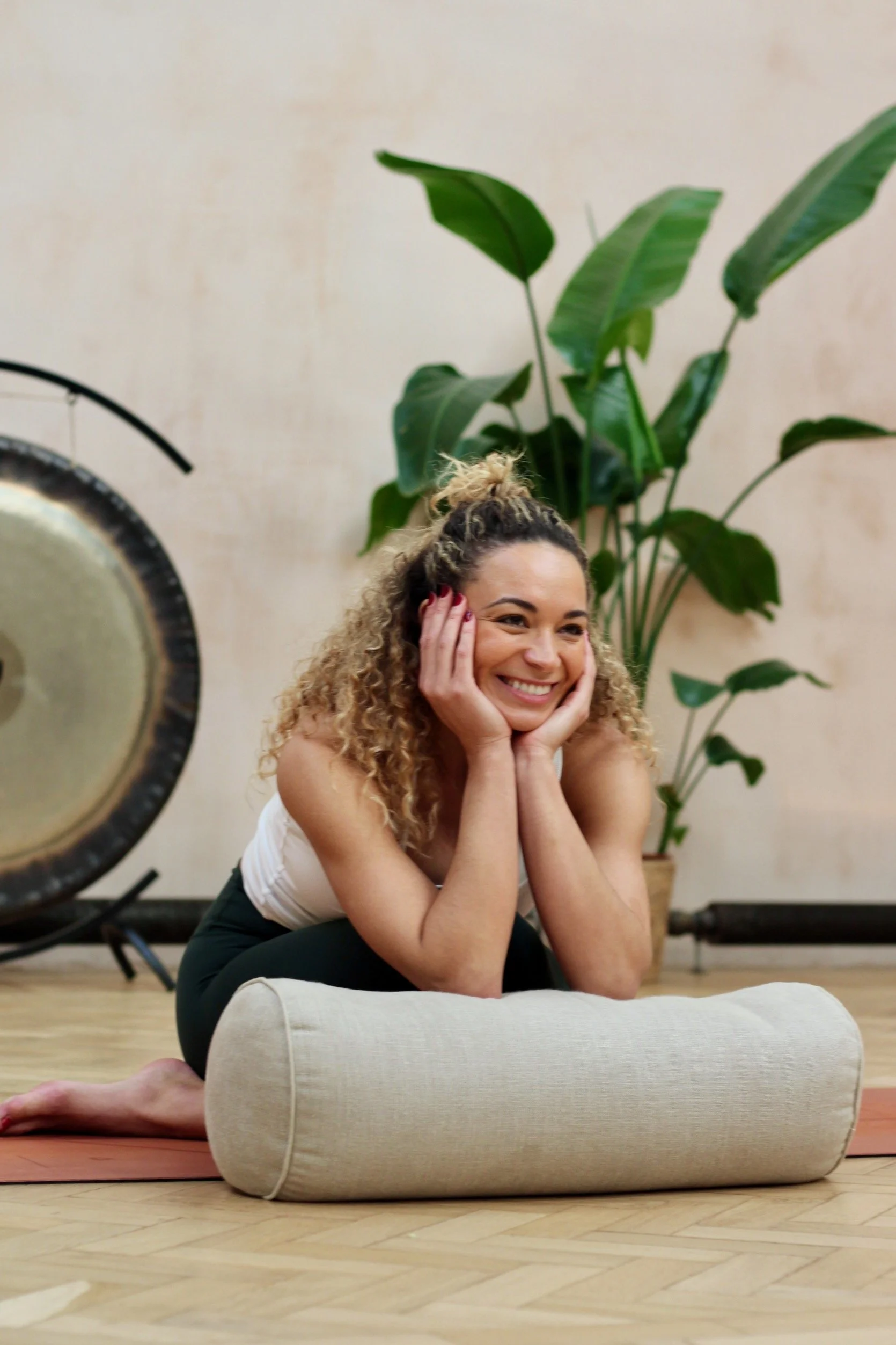 Woman smiling with curly hair, sitting on a yoga mat, supporting her chin with her hands, in front of a large plant, with a large studio lamp in the foreground and a beige wall in the background.