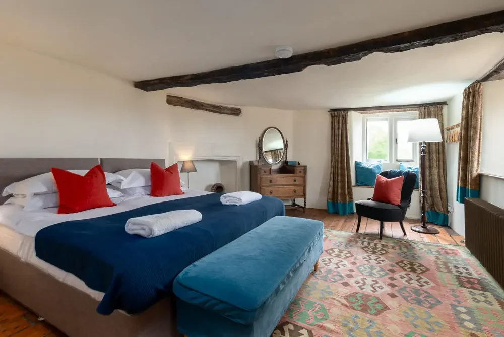 A cozy hotel room with a large bed that has white sheets, red pillows, and a dark blue bedspread, a small bedside lamp, a wooden dresser with a mirror, a sitting chair by the window with blue cushions, and a patterned rug on a wooden floor, with exposed wooden beams on the ceiling.
