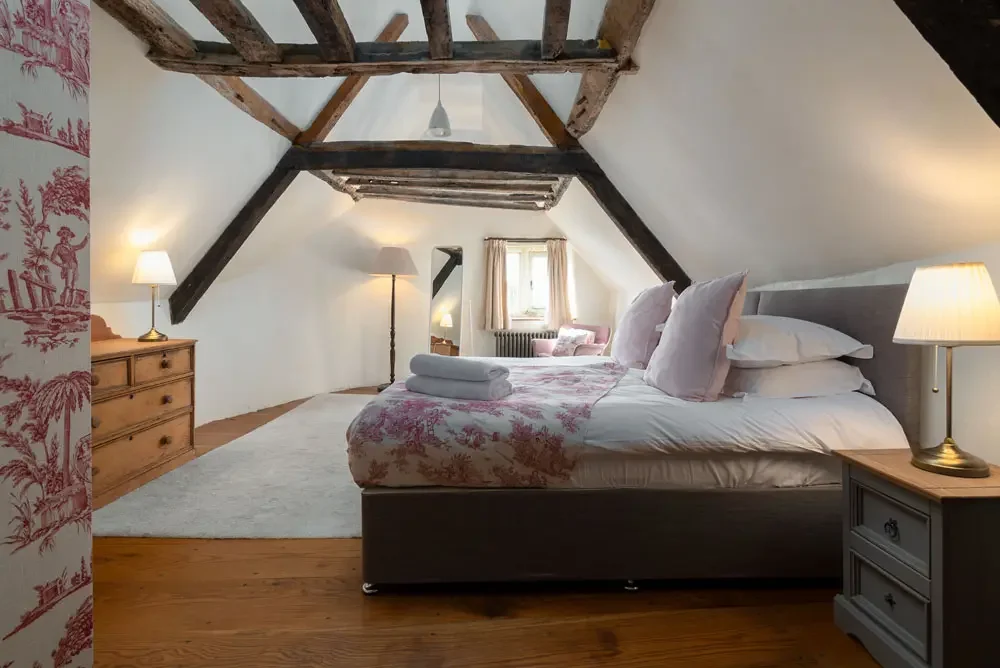 A cozy attic bedroom with a bed, bedside lamps, wooden dresser, and small seating area by the window, featuring exposed wooden beams and white walls.