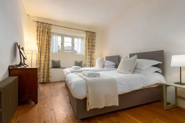 A bedroom with two beds share a nightstand, white bedding, and decorative pillows. A window with floral curtains is in the background, and there is a chair with cushions by the window. The room has wooden floors and minimal decoration.