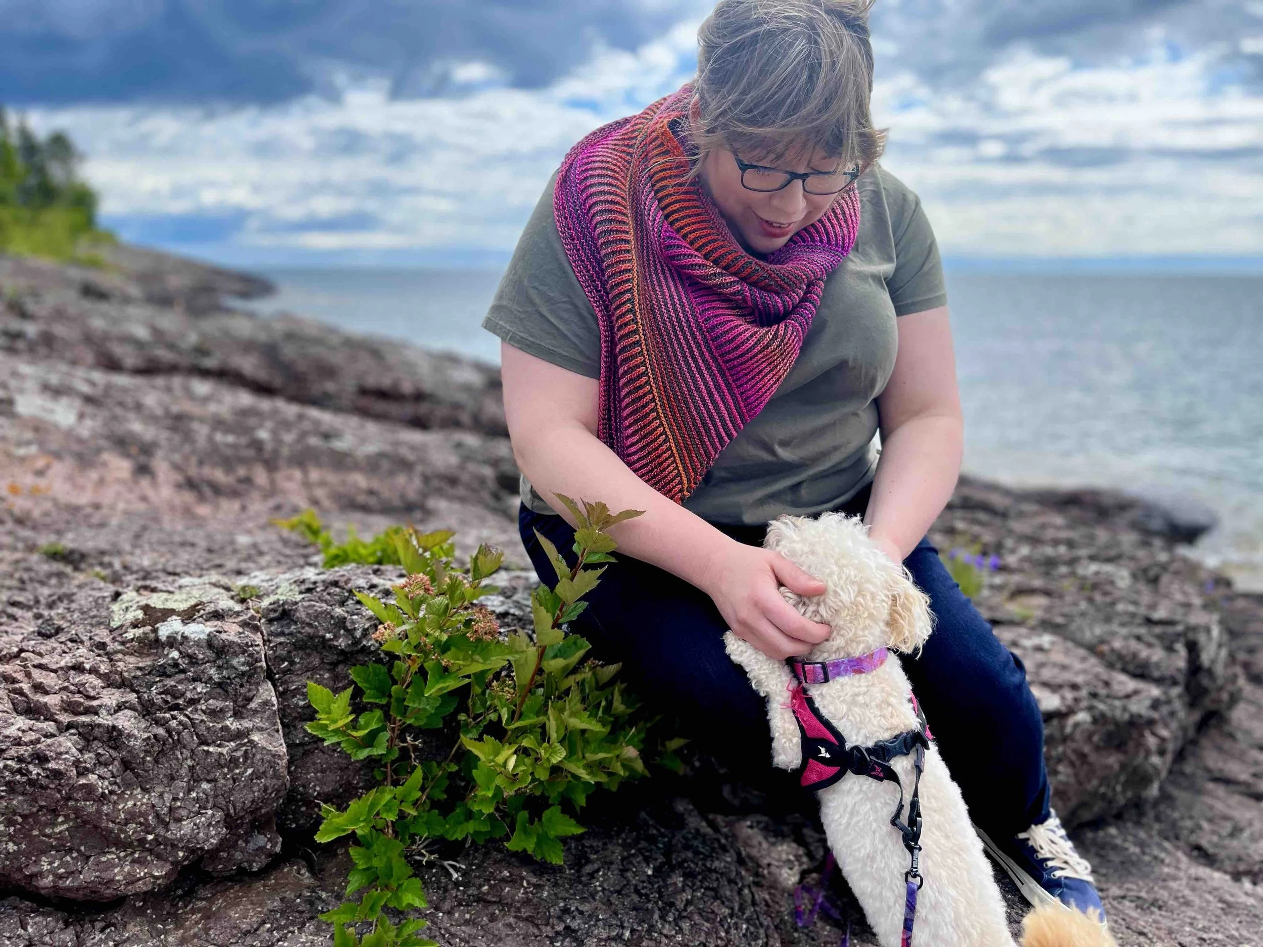 Katie, a white woman with light brown hair, leans down to pet the ears of a small cream poodle. Around her neck is a bright pink and olive green Montrose cowl