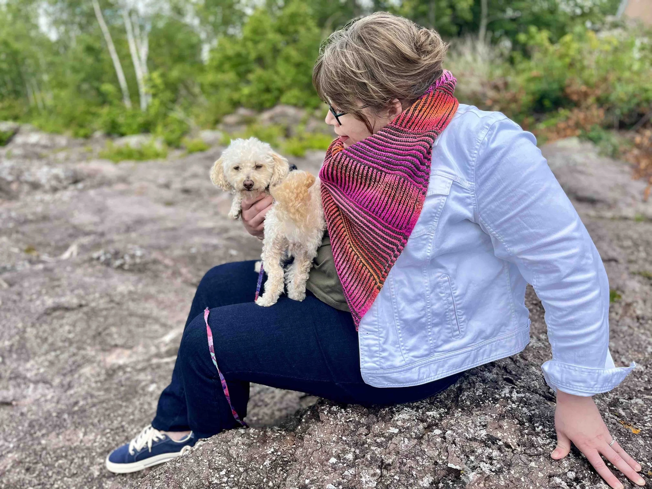 A woman, wearing a bright pink striped montrose cowl over a white jean jacket, blue jeans, and blue tennis shoes, holds a small cream colored poodle while sitting on a rocky shoreline