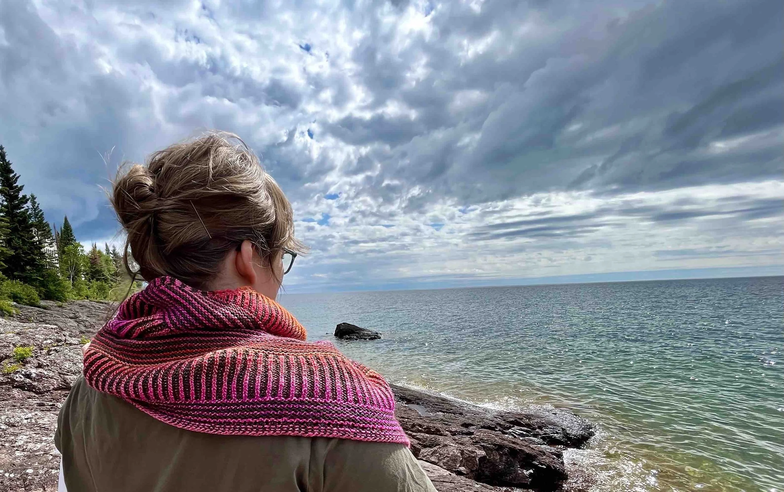 A white woman with brown hair looks out at Lake Superior. Around her shoulders is a bright pink and olive green striped montrose cowl