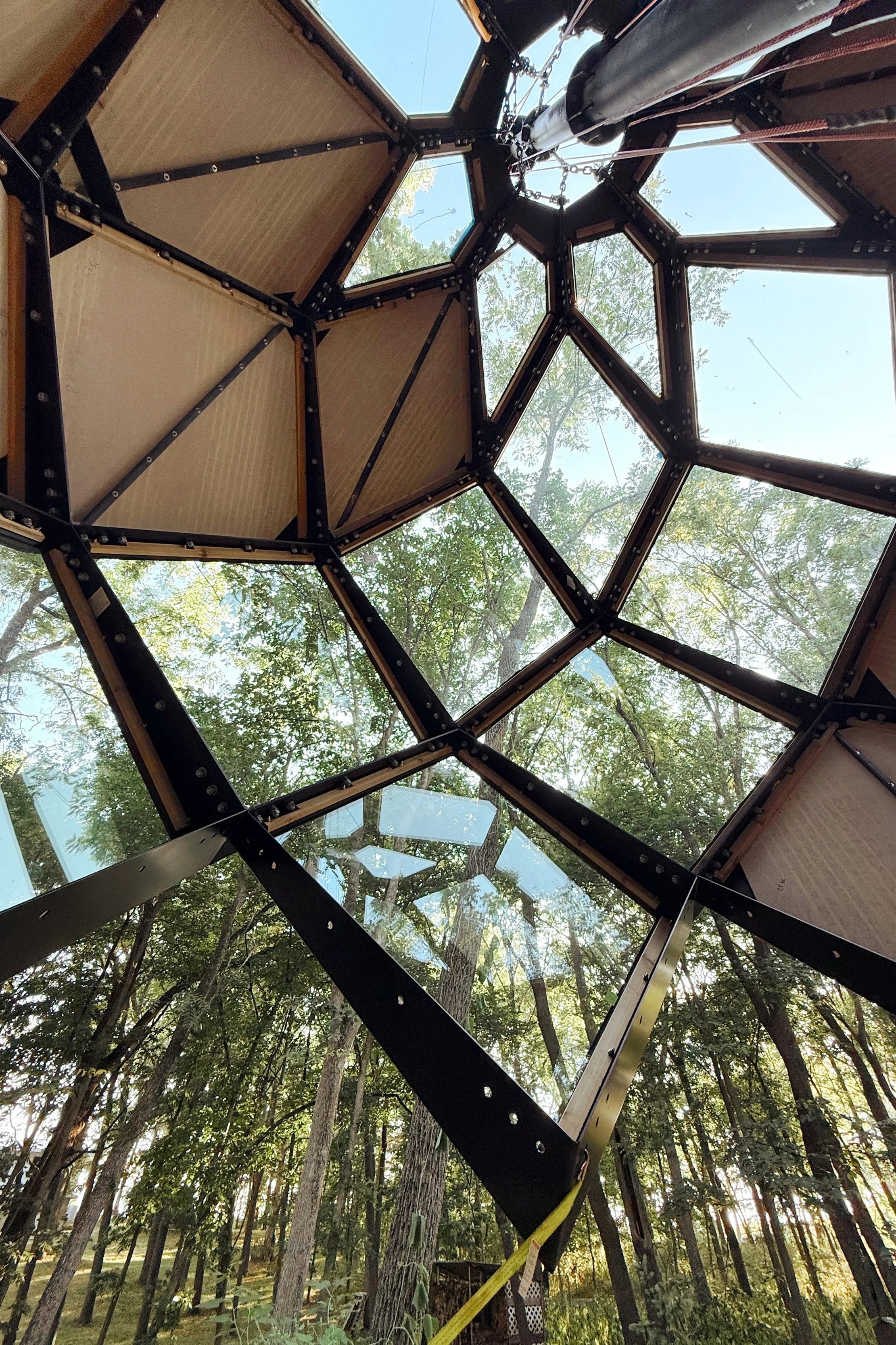 View of the underside of a geodesic dome made up of hexagonal and pentagonal glass panels with a support structure of black metal beams, set against trees and sky outside.