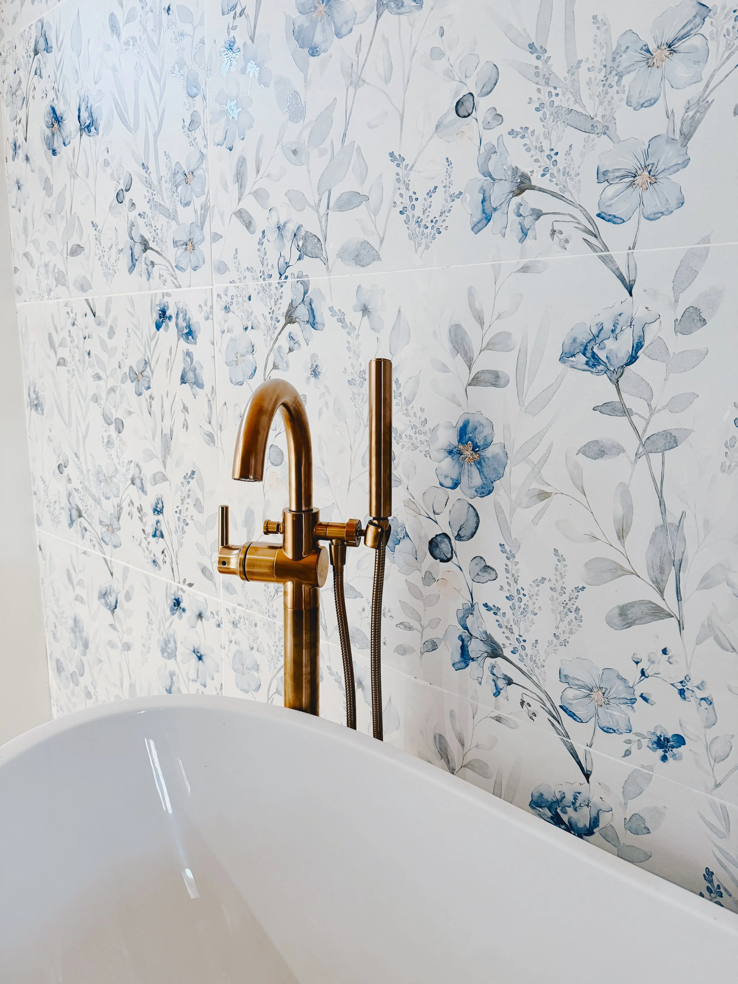 Close-up of a bathroom bathtub with a gold-colored faucet and a blue-flower patterned tiled wall in the background.