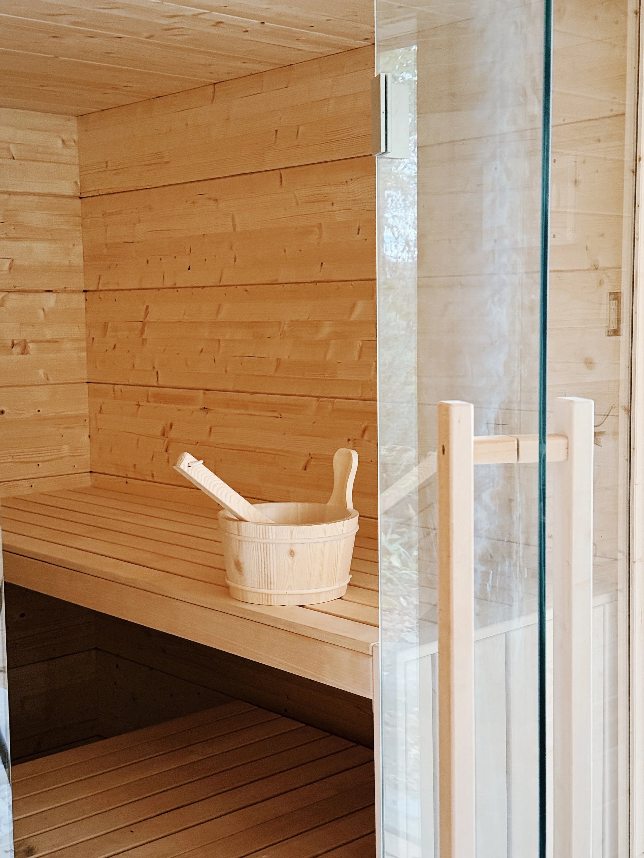 Interior of a wooden sauna room with a wooden bucket and ladle on a bench, and a glass door with wooden handles leading into the sauna.