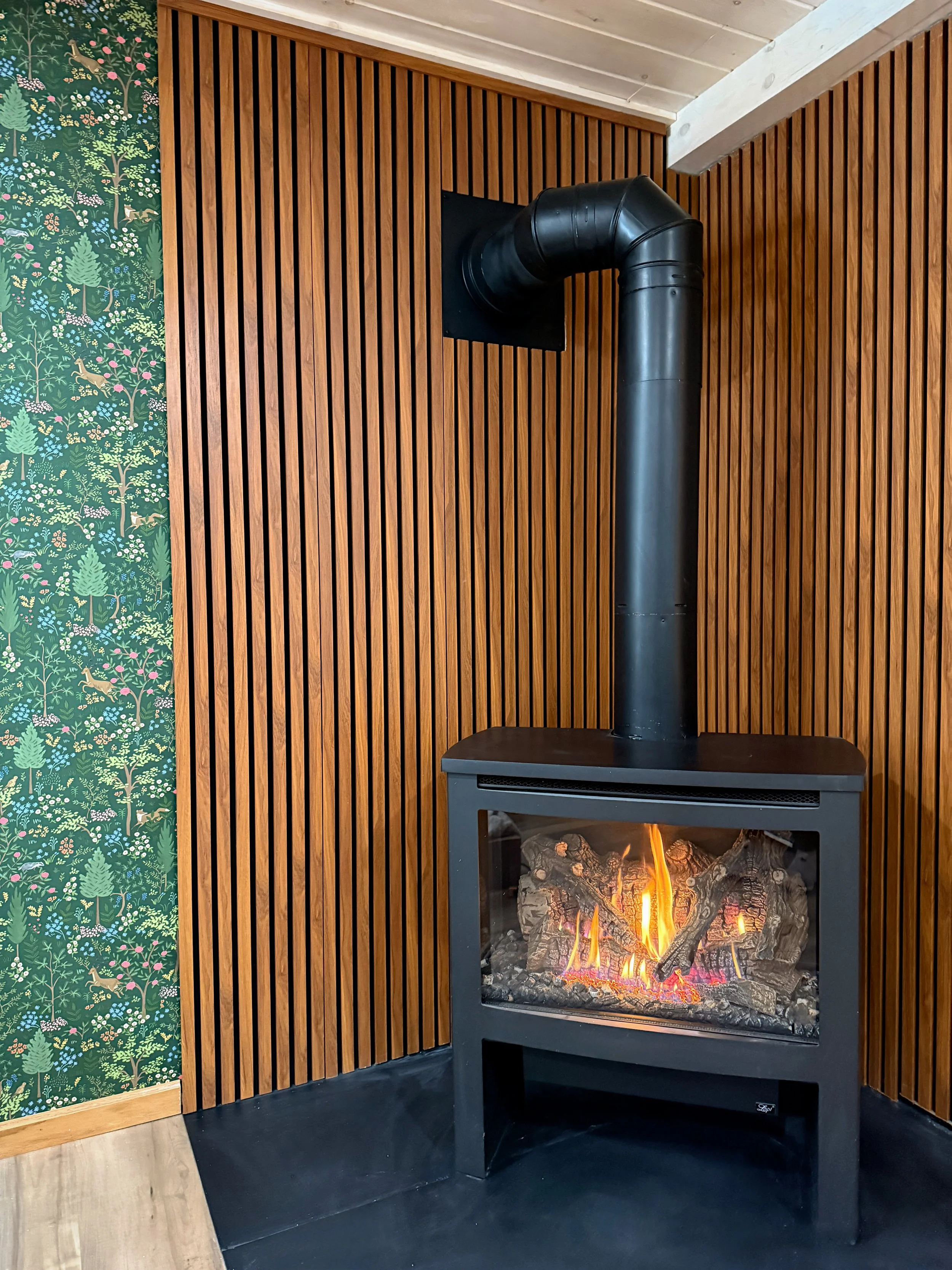 A modern black wood stove with a glass front showing a burning fire with logs inside, installed against a wood-paneled wall with vertical slats, and a corner of a floral patterned wallpaper on the left. The stove's ventilation pipe extends from the top into the wall.