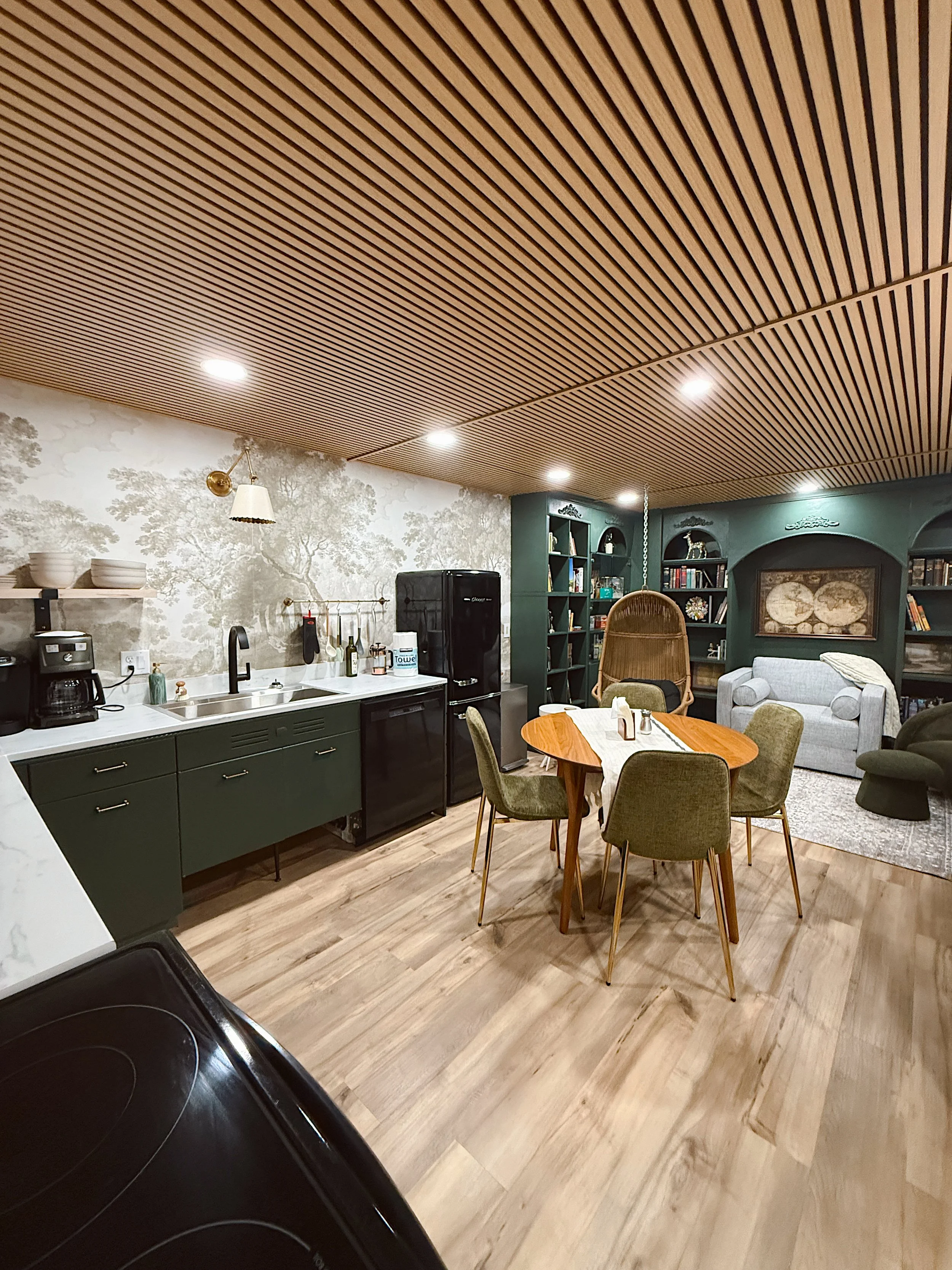 Interior of a cozy living space featuring a kitchenette with green cabinets, a round wooden dining table with six chairs, shelves filled with books, a white sofa, an armchair, and a wall with a mural of trees. The ceiling has wooden slats and recessed lighting.