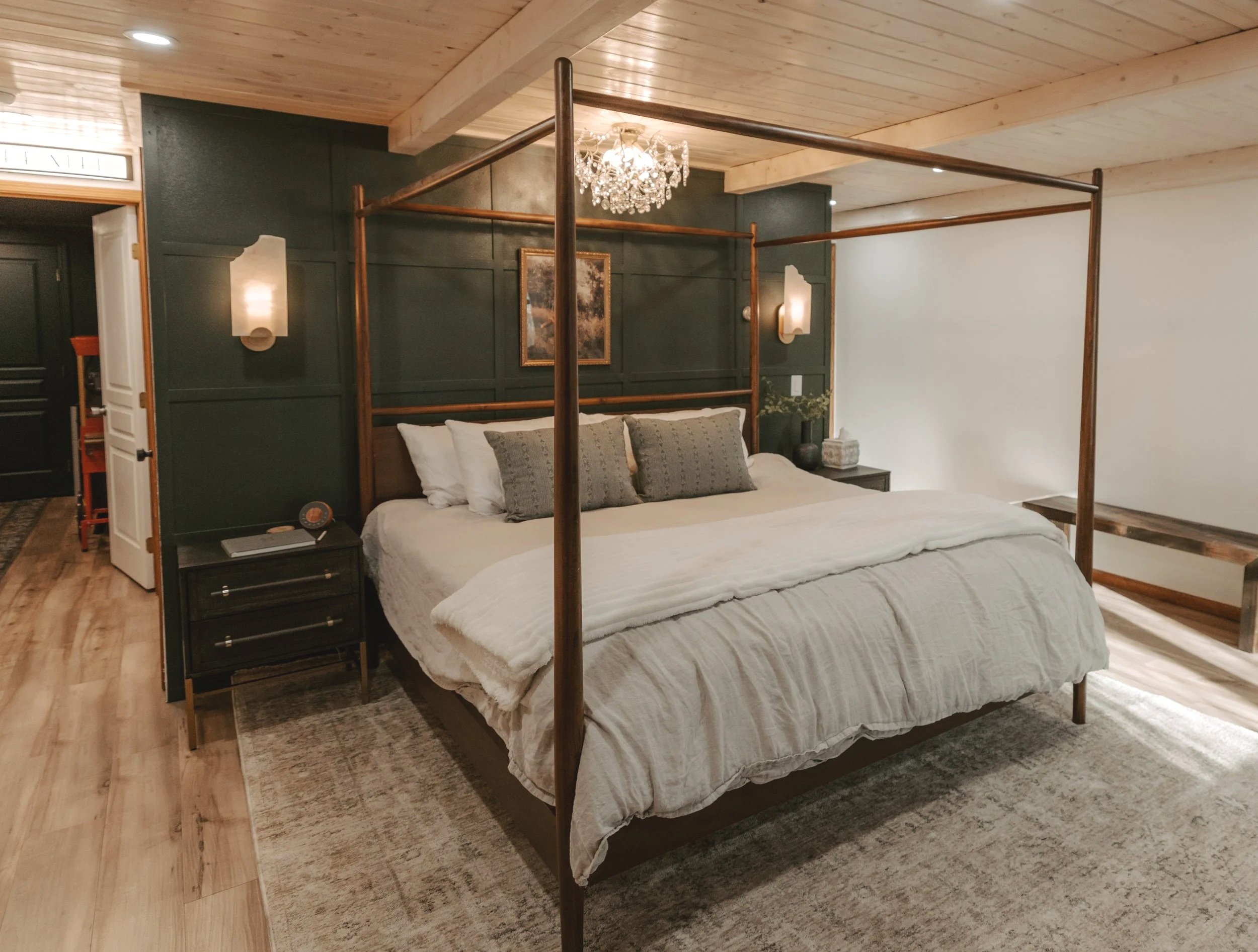 A bedroom with a wooden four-poster bed with white bedding and green pillows, dark green accent wall, wood nightstands, wall-mounted lamps, and a small chandelier.