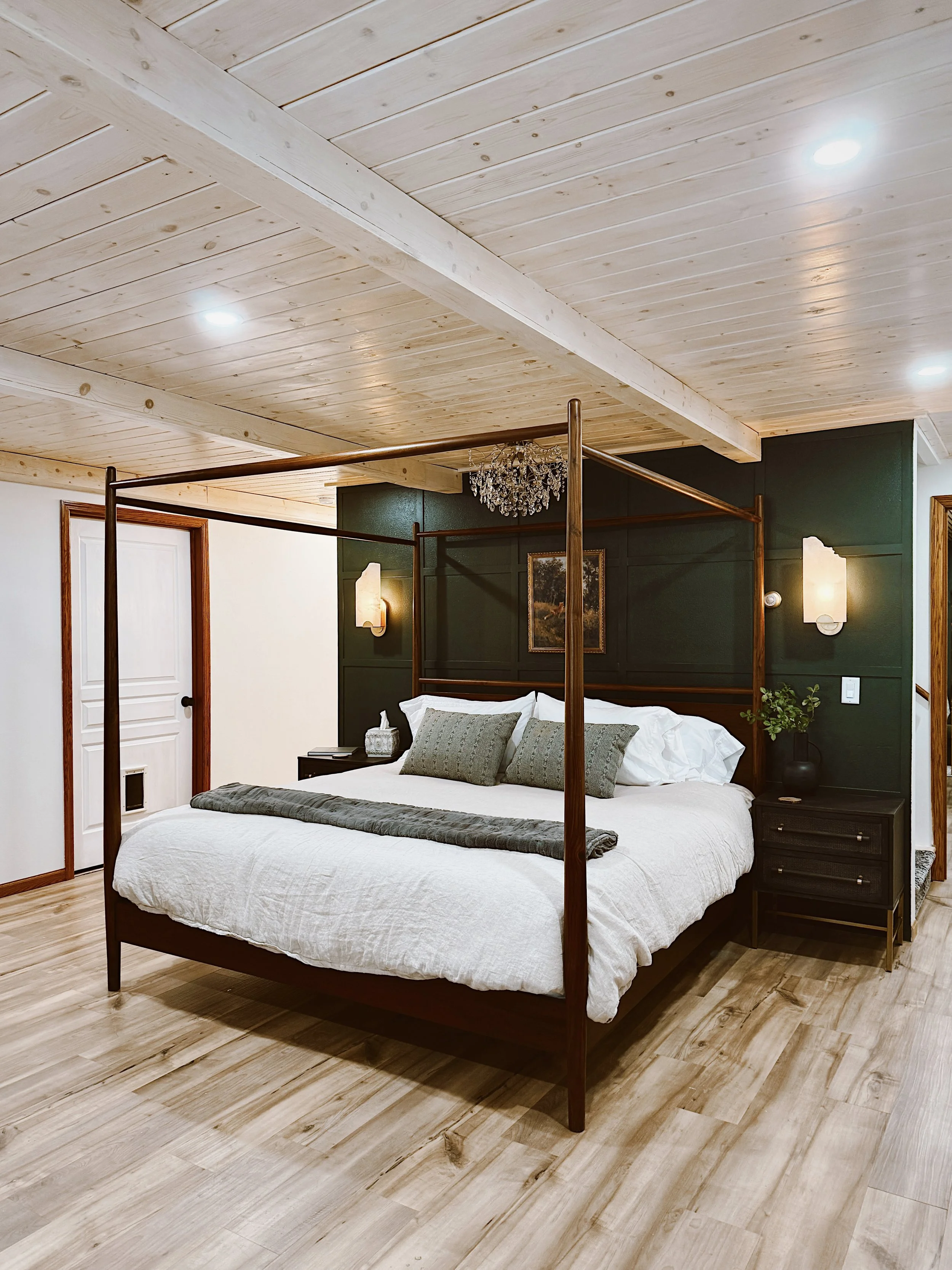 A bedroom with a wooden four-poster bed with white bedding and green pillows, dark green accent wall, wood nightstands, wall-mounted lamps, and a small chandelier.