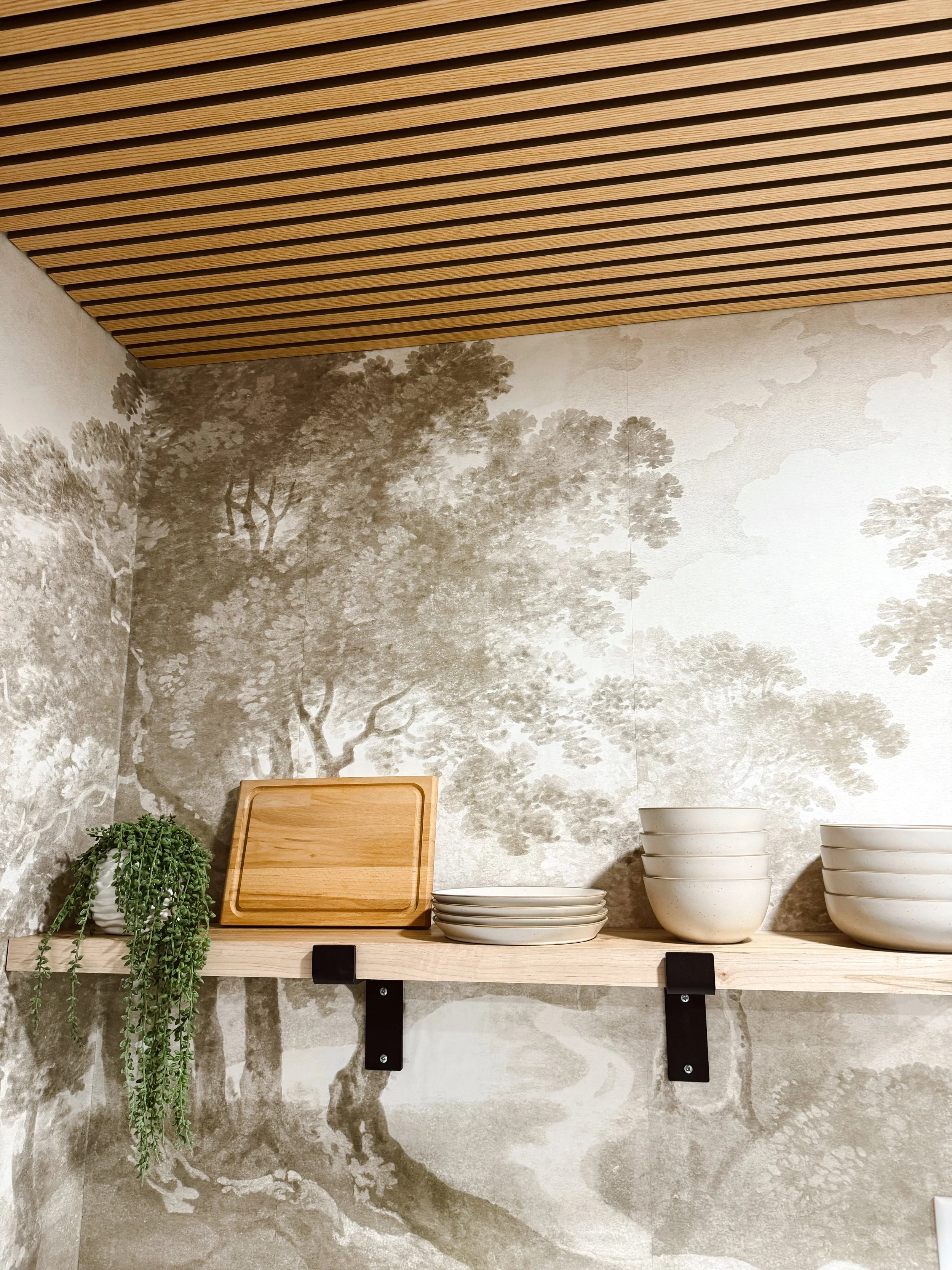 A wooden shelf with a potted plant, a wooden cutting board, strawberry-shaped plates, and bowls against a wall with mural of trees and clouds.  The ceiling has wooden slats.