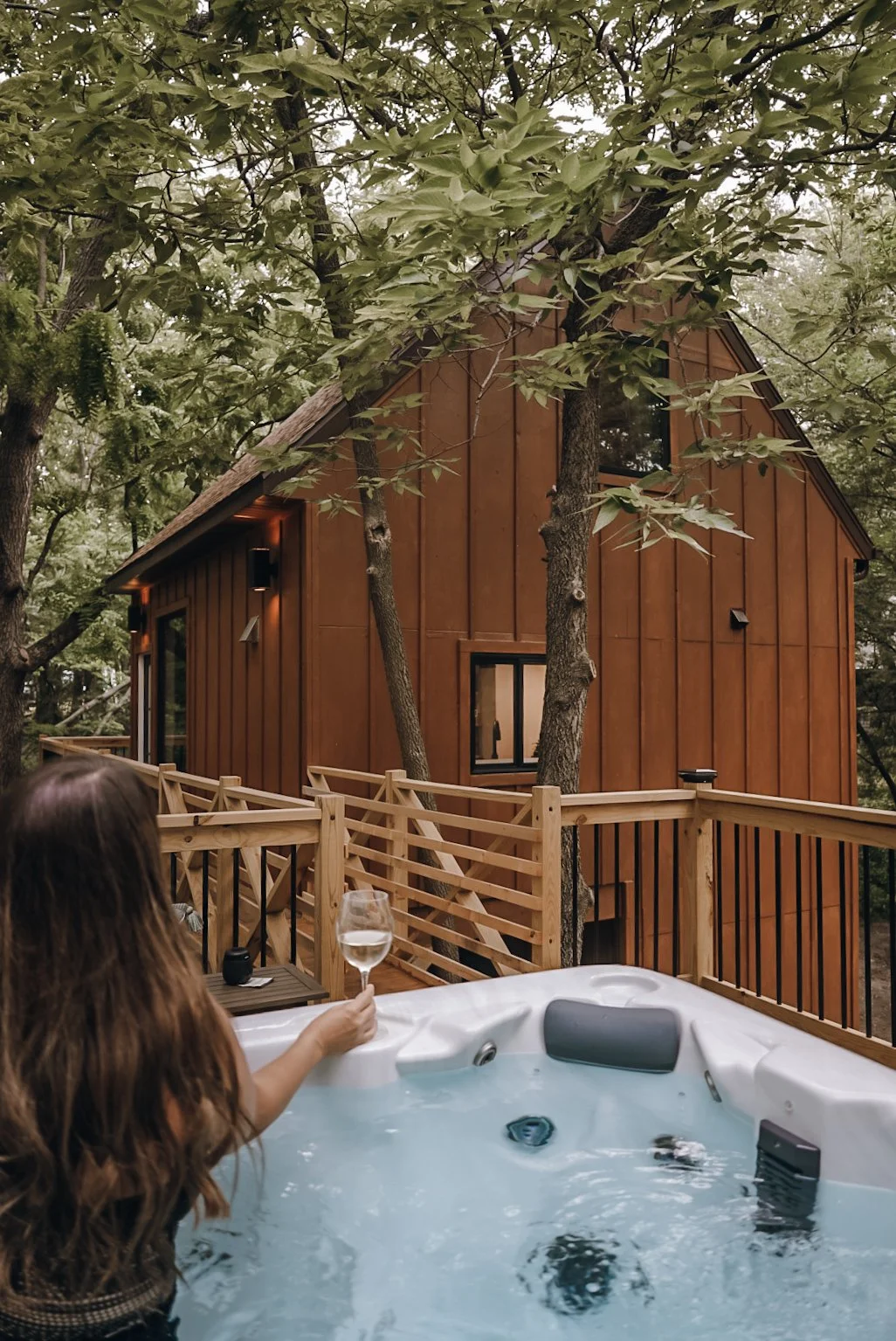 A woman relaxing in a hot tub on a wooden deck surrounded by trees, holding a glass of white wine, with a brown house with large windows in the background.