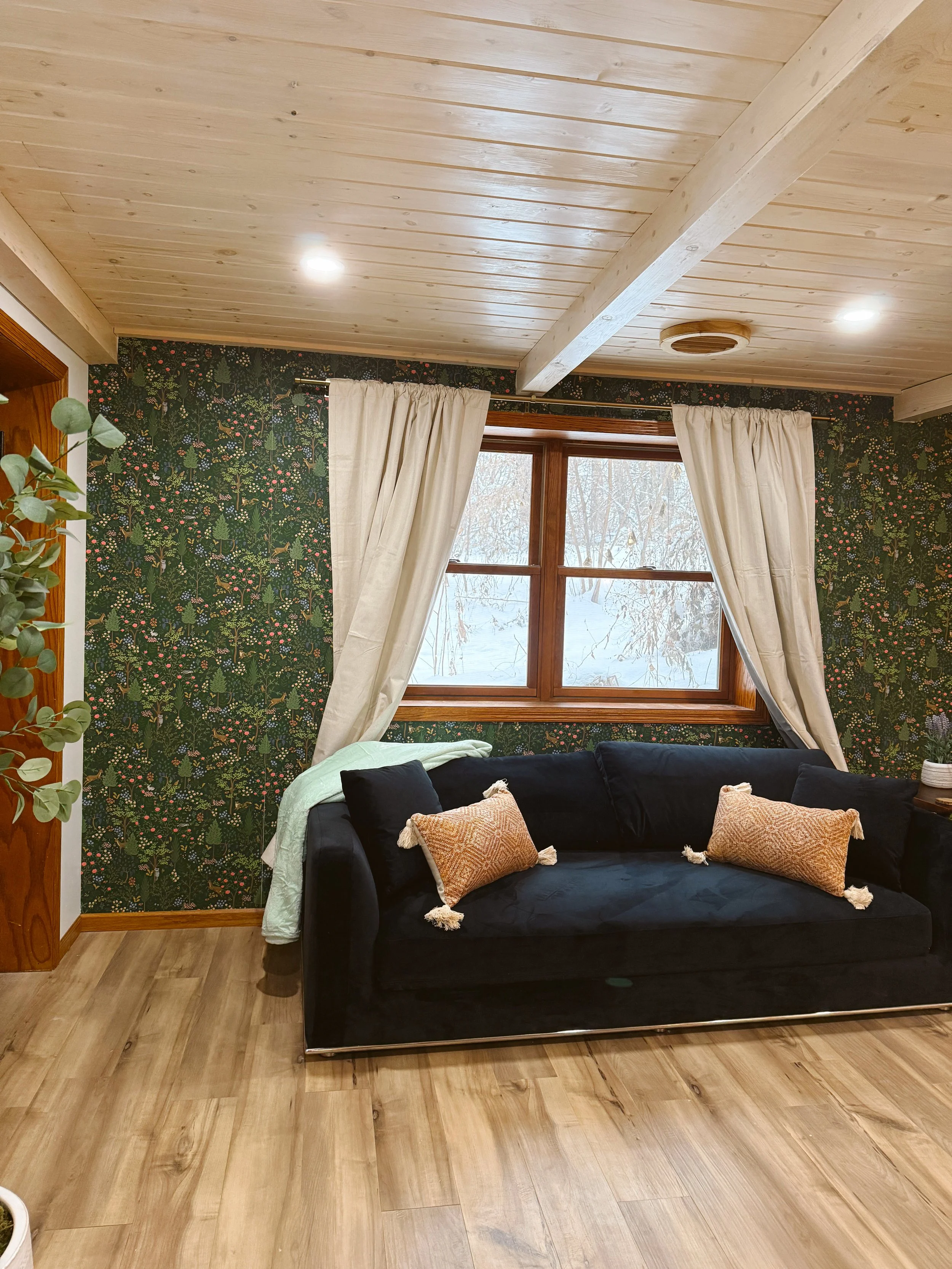 Living room with black velvet sofa, beige throw pillows with tassels, white curtains, wooden window, floral patterned wallpaper, wooden ceiling with recessed lighting, wooden flooring, and a potted plant on the left side.