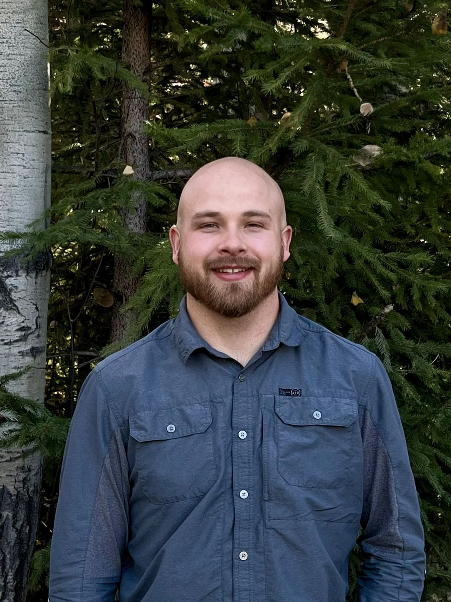 A man with a beard and no hair smiling outdoors in front of a background of evergreen trees and a birch tree.