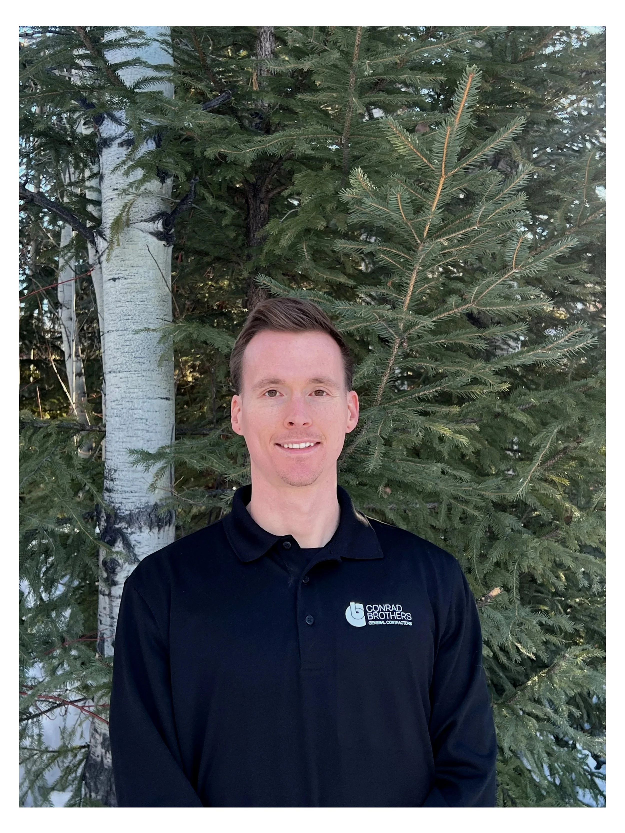 A man with short brown hair smiling, wearing a black polo shirt with a logo that reads 'Conrad Brothers General Contractors,' standing outdoors in front of a large pine tree with snow on the ground.