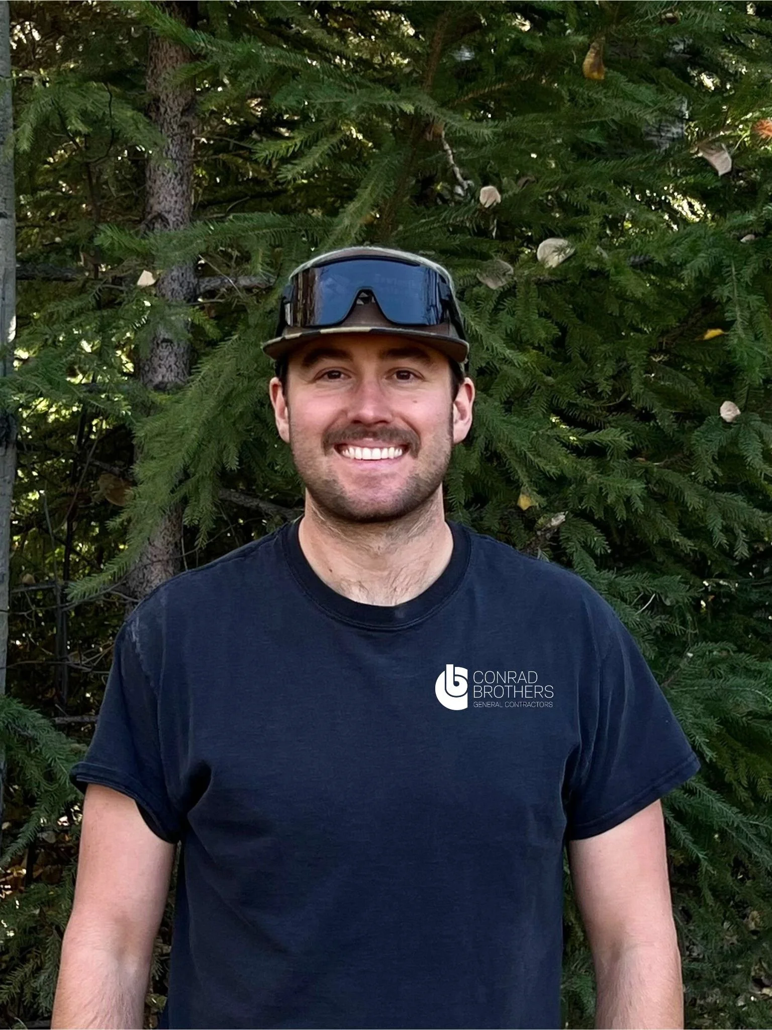 A smiling man wearing a black T-shirt with 'Conrad Brothers General Contractors' logo, standing outdoors in front of a lush green pine tree, with sunglasses on his head.