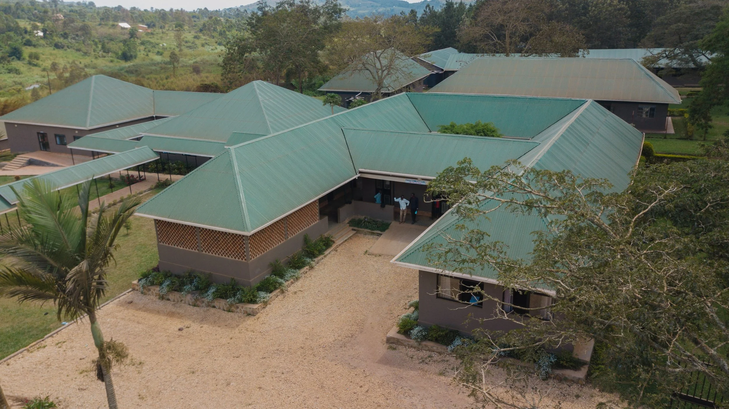 An aerial view of several buildings with green metal roofs, surrounded by trees and a landscape, with a few people standing at the entrance.