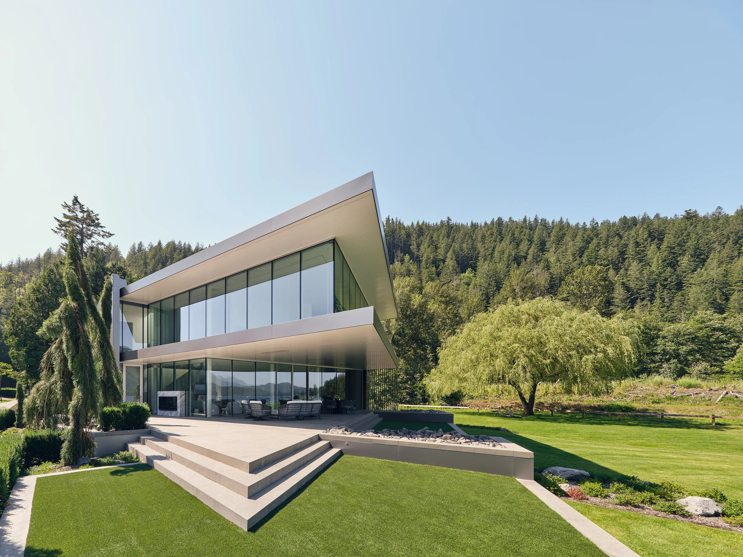 Modern house with glass walls and flat roof, surrounded by green lawn, trees, and forested hills under a clear blue sky.